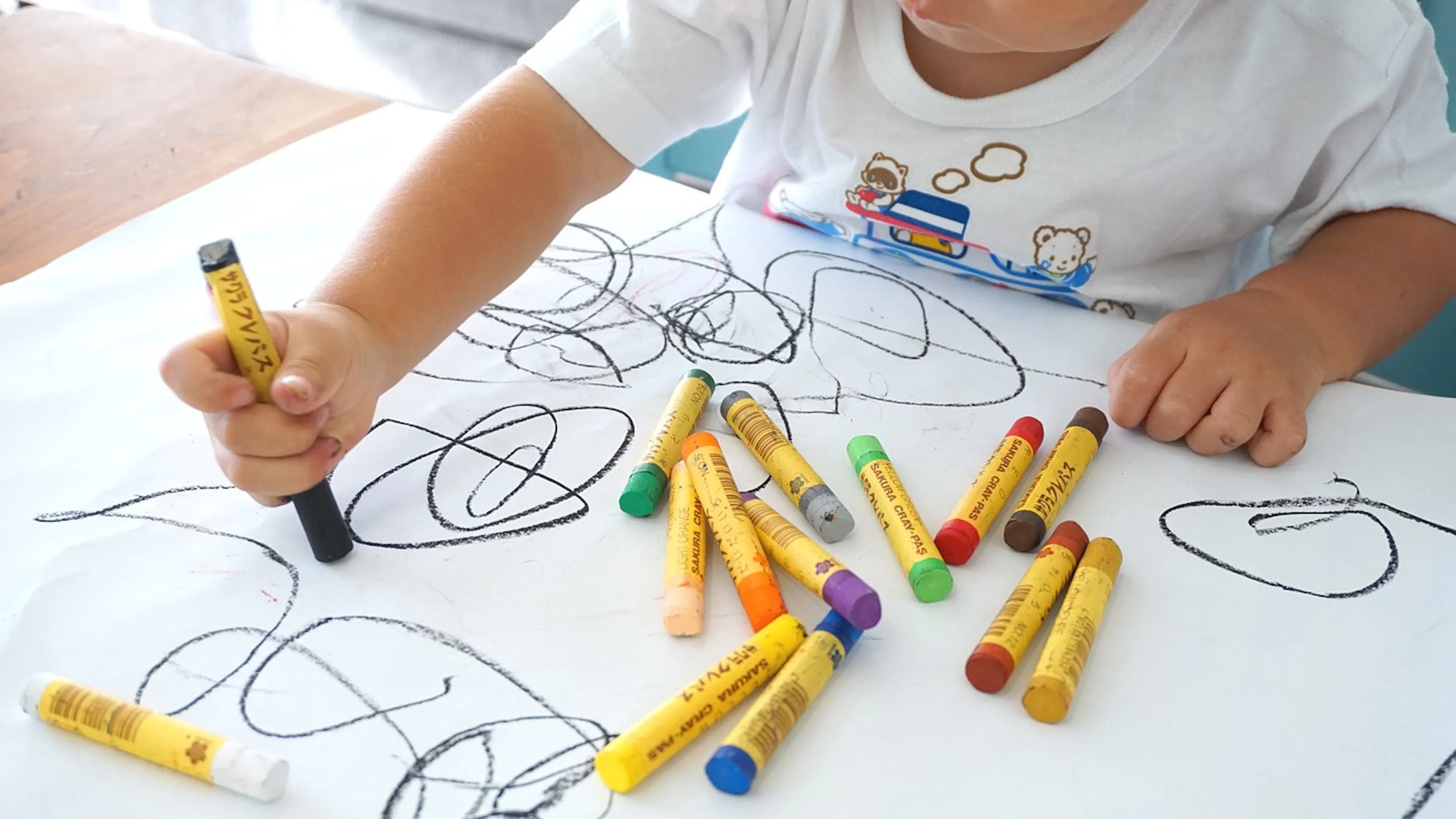 A baby drawing squiggles with some multicolored crayons on a big white sheet of paper