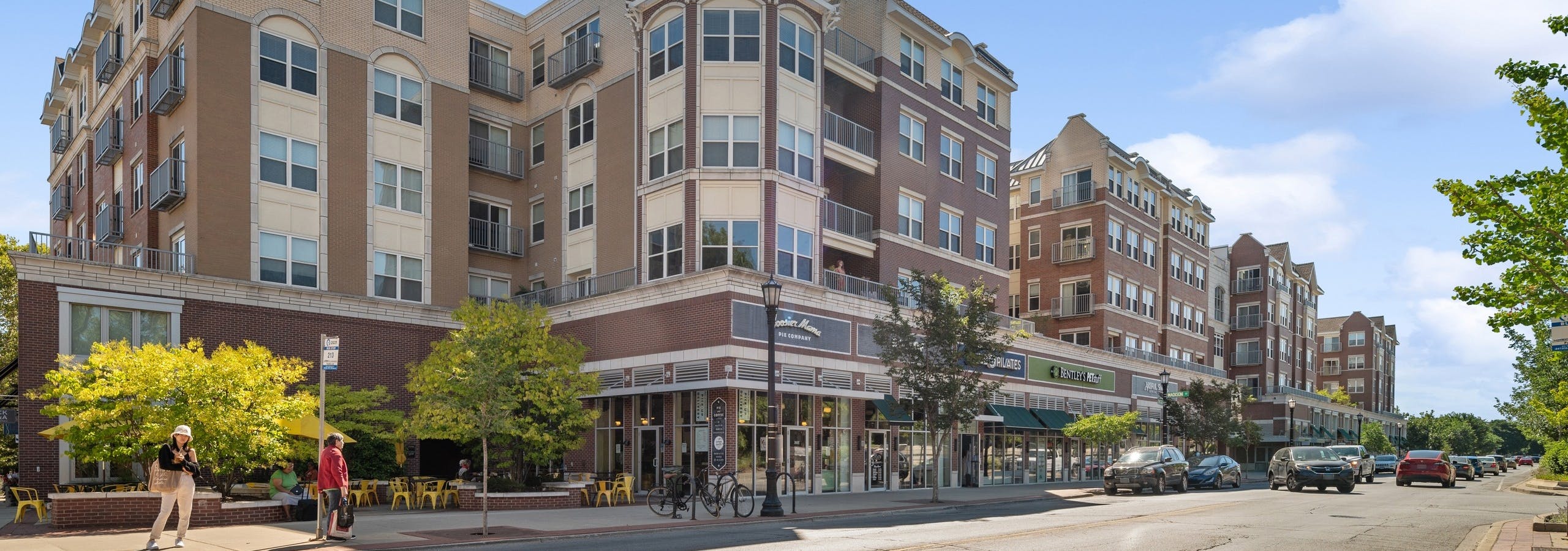 Street corner view of AMLI Evanston apartment building brick façade with retail stores and cars and pedestrians under blue sky