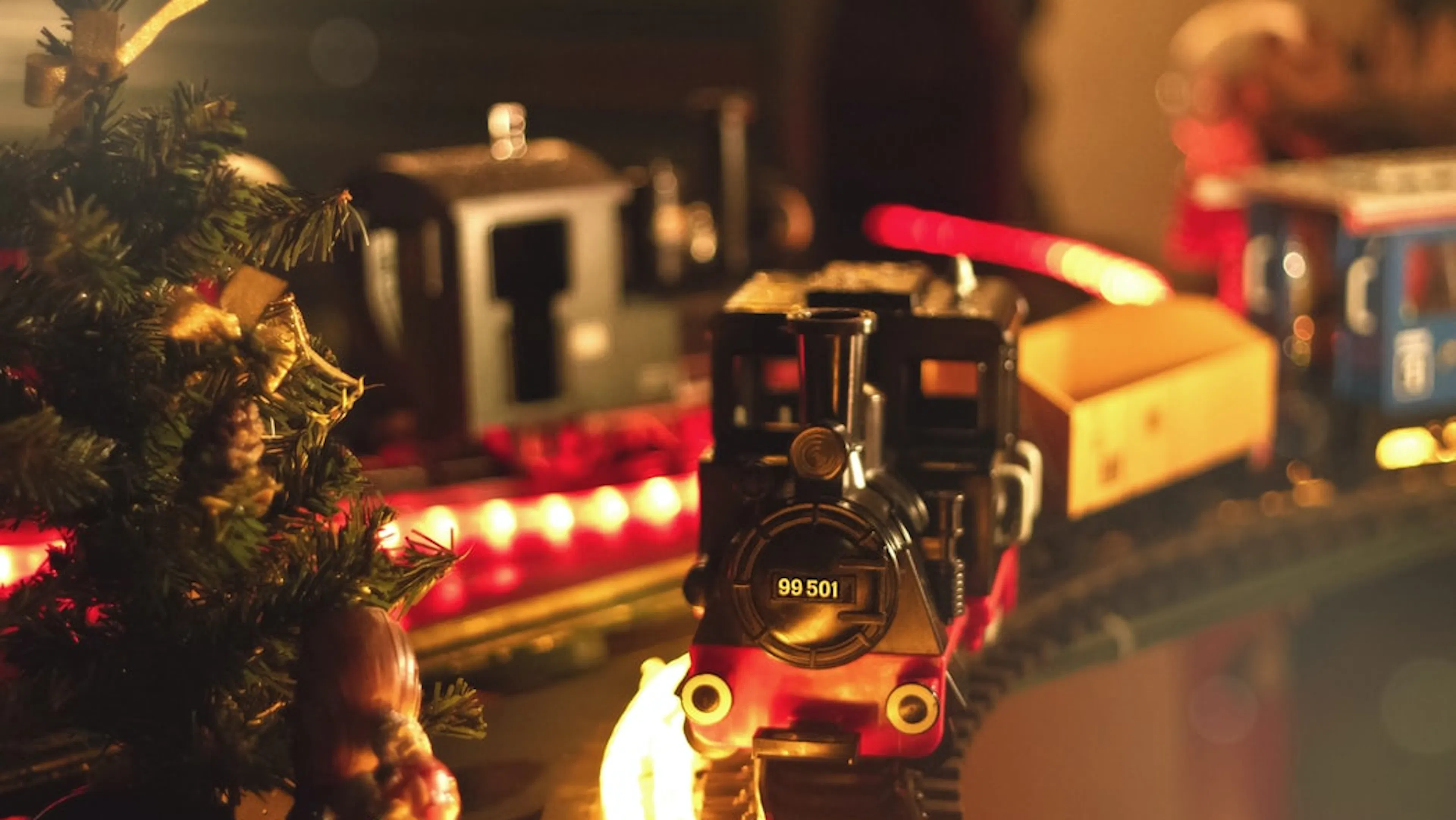 A black and red toy train on a toy train track passing by a small Christmas tree dimly lit by string lights