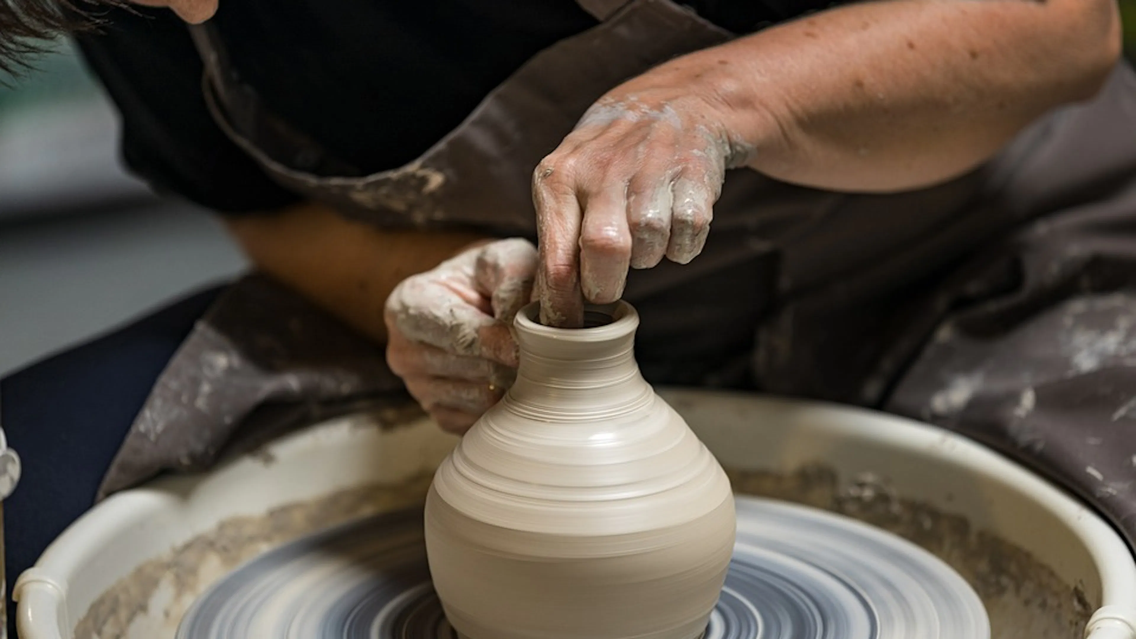 A woman crouching over a potters wheel and spinning a clay vase