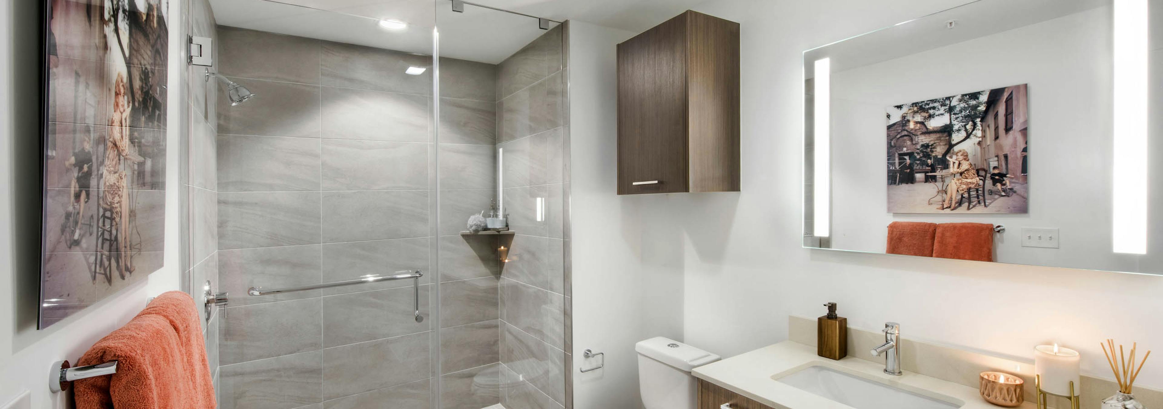 Bathroom at AMLI Midtown 29 with glass enclosed tile shower and wood finish cabinets with white countertops and lighted mirror