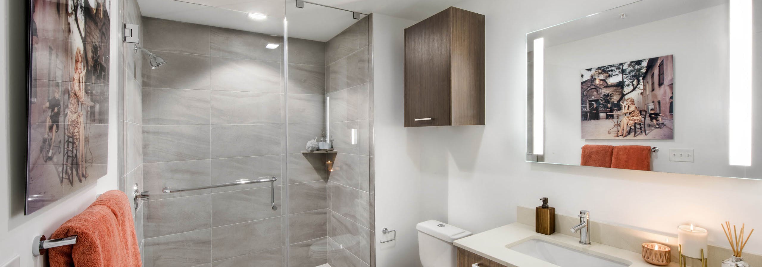 Bathroom at AMLI Midtown 29 with glass enclosed tile shower and wood finish cabinets with white countertops and lighted mirror