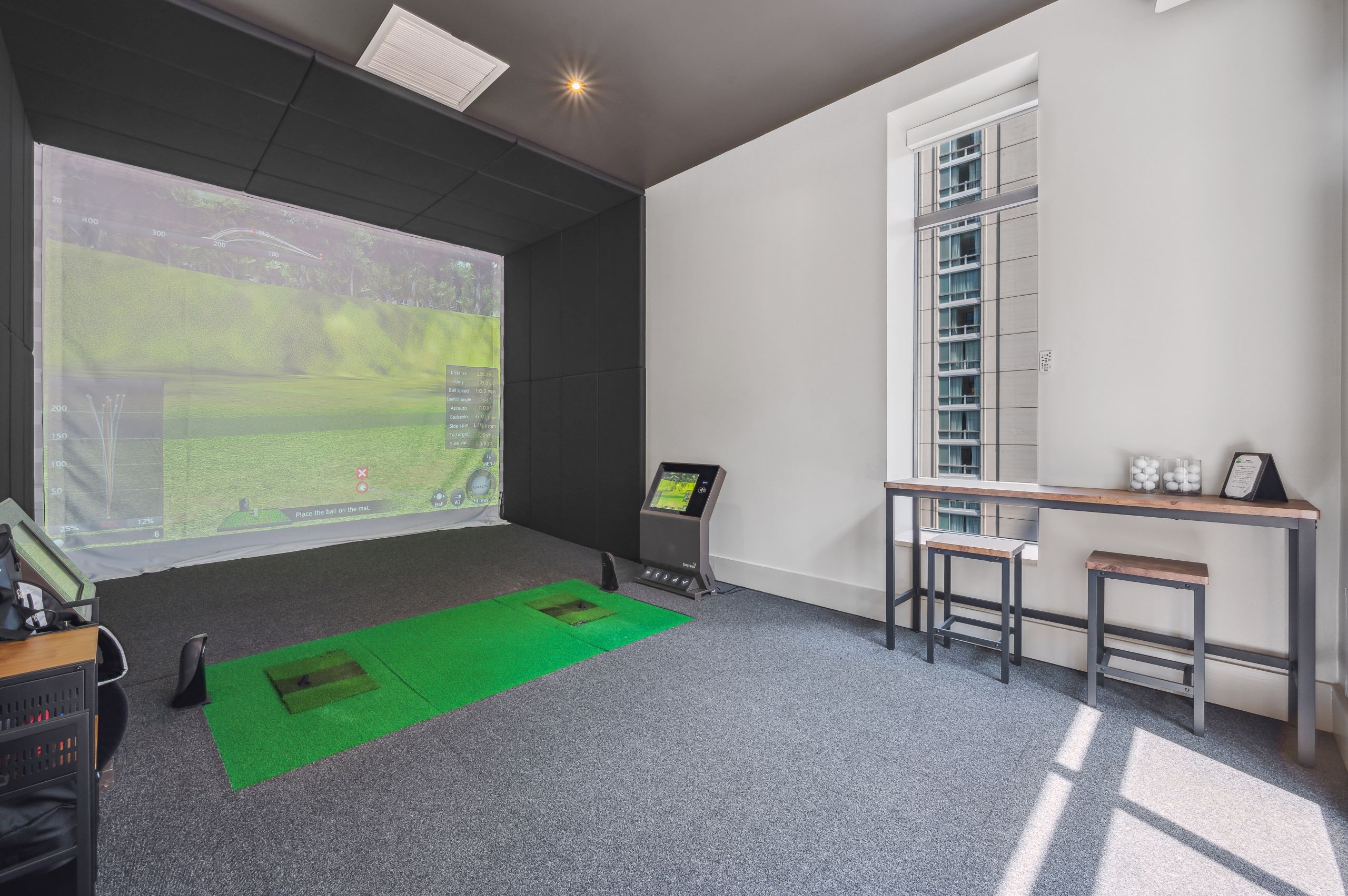 Golf simulator at AMLI River North with large screen with golf scene and a wood table with 2 stools with gray carpeting 