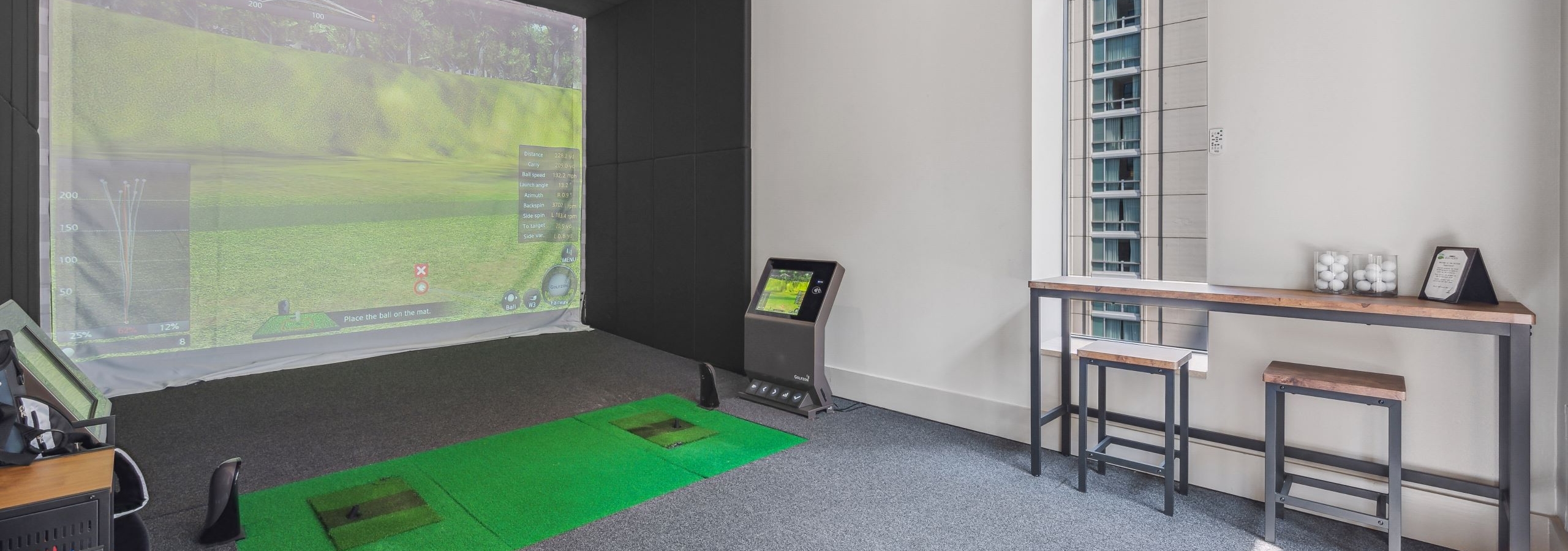Golf simulator at AMLI River North with large screen with golf scene and a wood table with 2 stools with gray carpeting