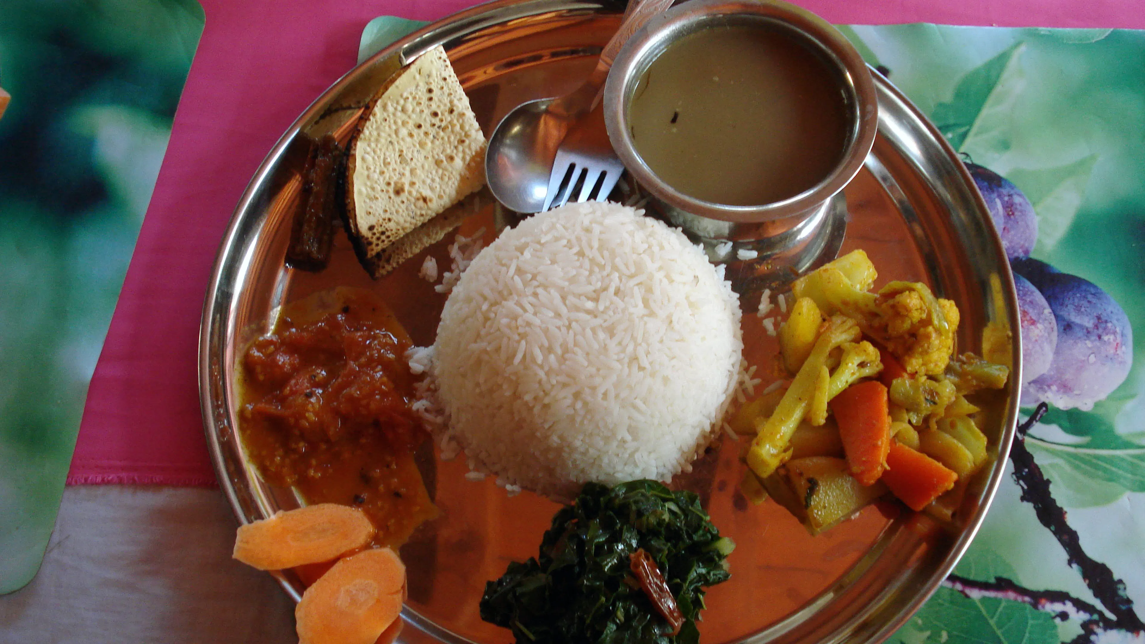 Metal plate with a mound of rice, a bowl of lentil soup, some green vegetables and a corn chip