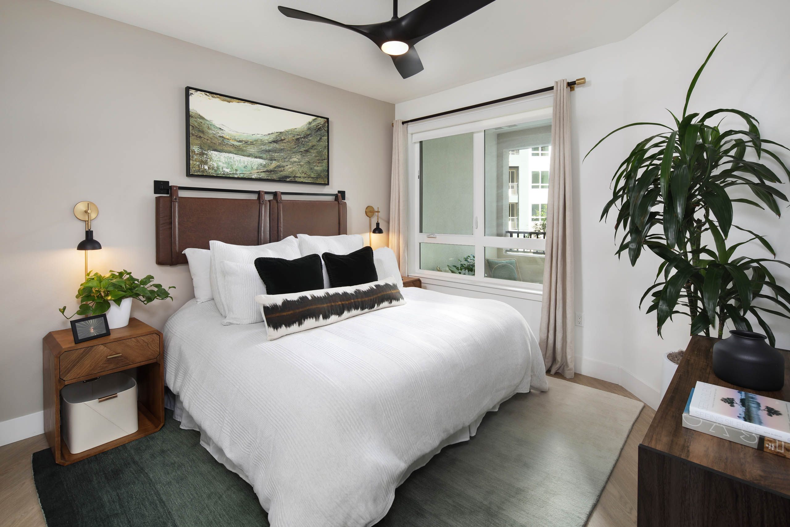 Bedroom with white bedding, leather-style headboard, ceiling fan, landscape artwork, nightstands, large window with curtains, and tall plant.