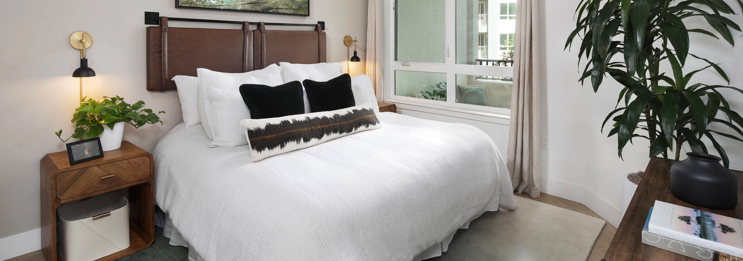 Bedroom with white bedding, leather-style headboard, ceiling fan, landscape artwork, nightstands, large window with curtains, and tall plant.