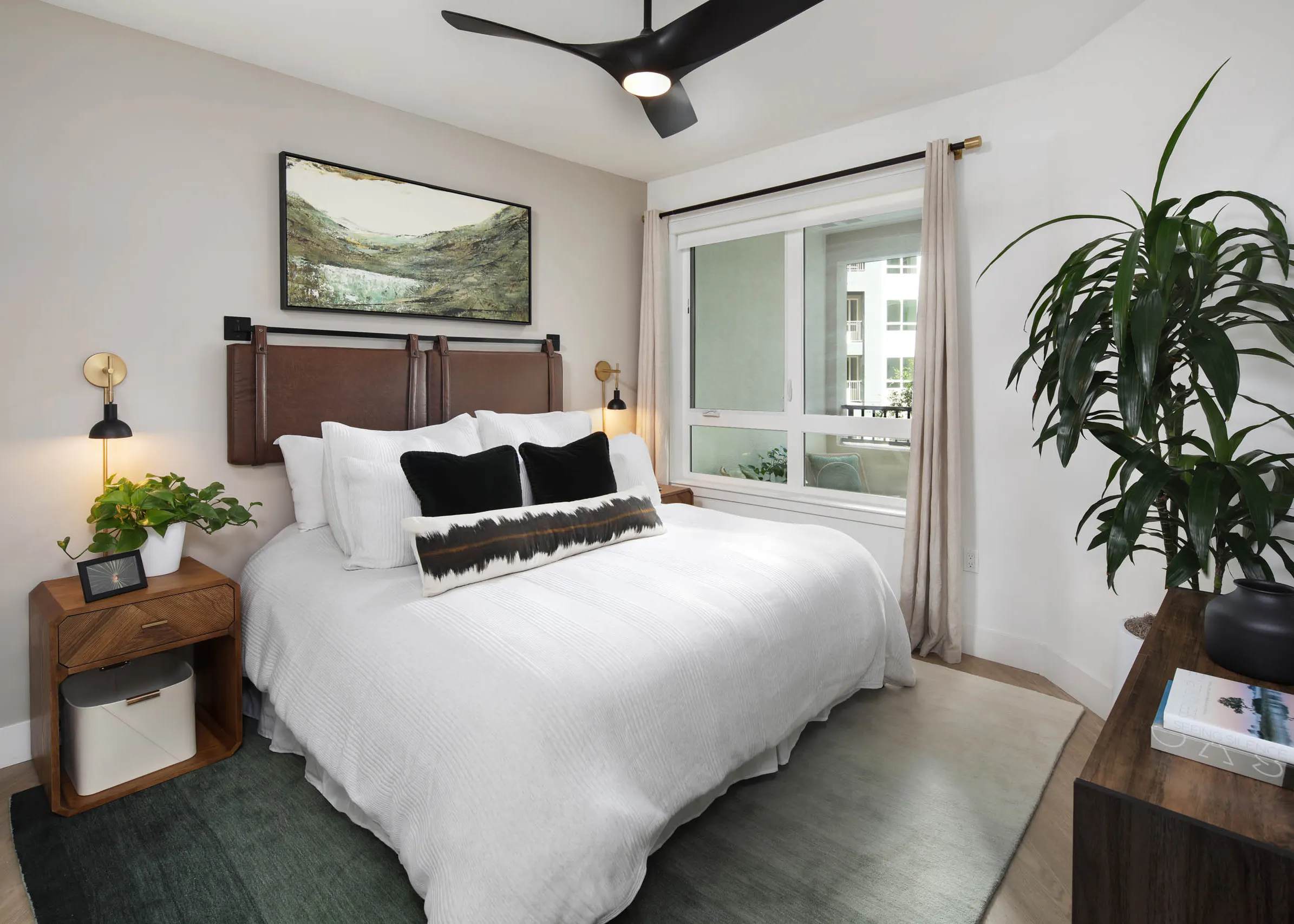 Bedroom with white bedding, leather-style headboard, ceiling fan, landscape artwork, nightstands, large window with curtains, and tall plant.