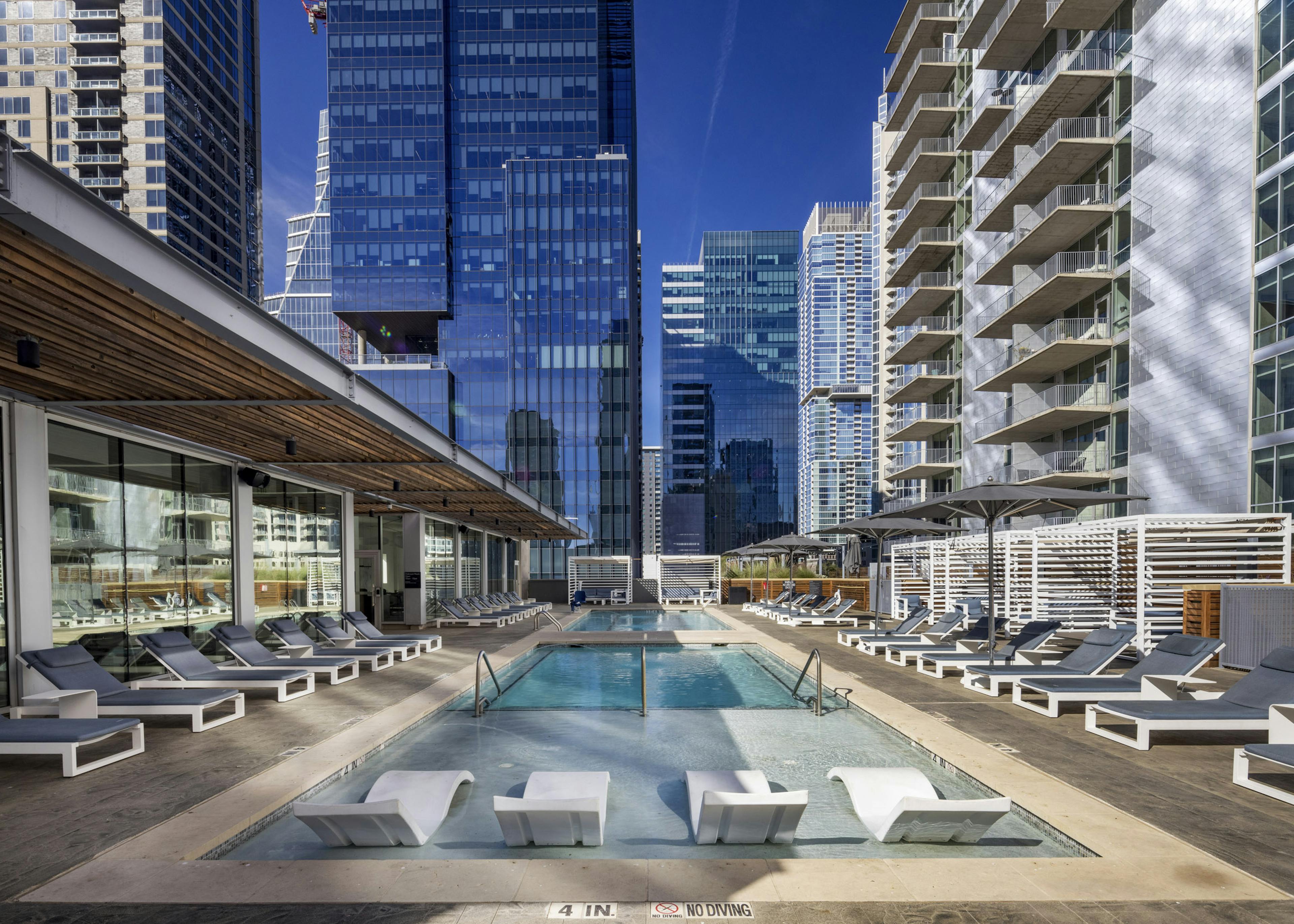Close up view of sleek white lounge chairs in shallow portion of AMLI on 2nd rooftop pool surrounded by daytime city views