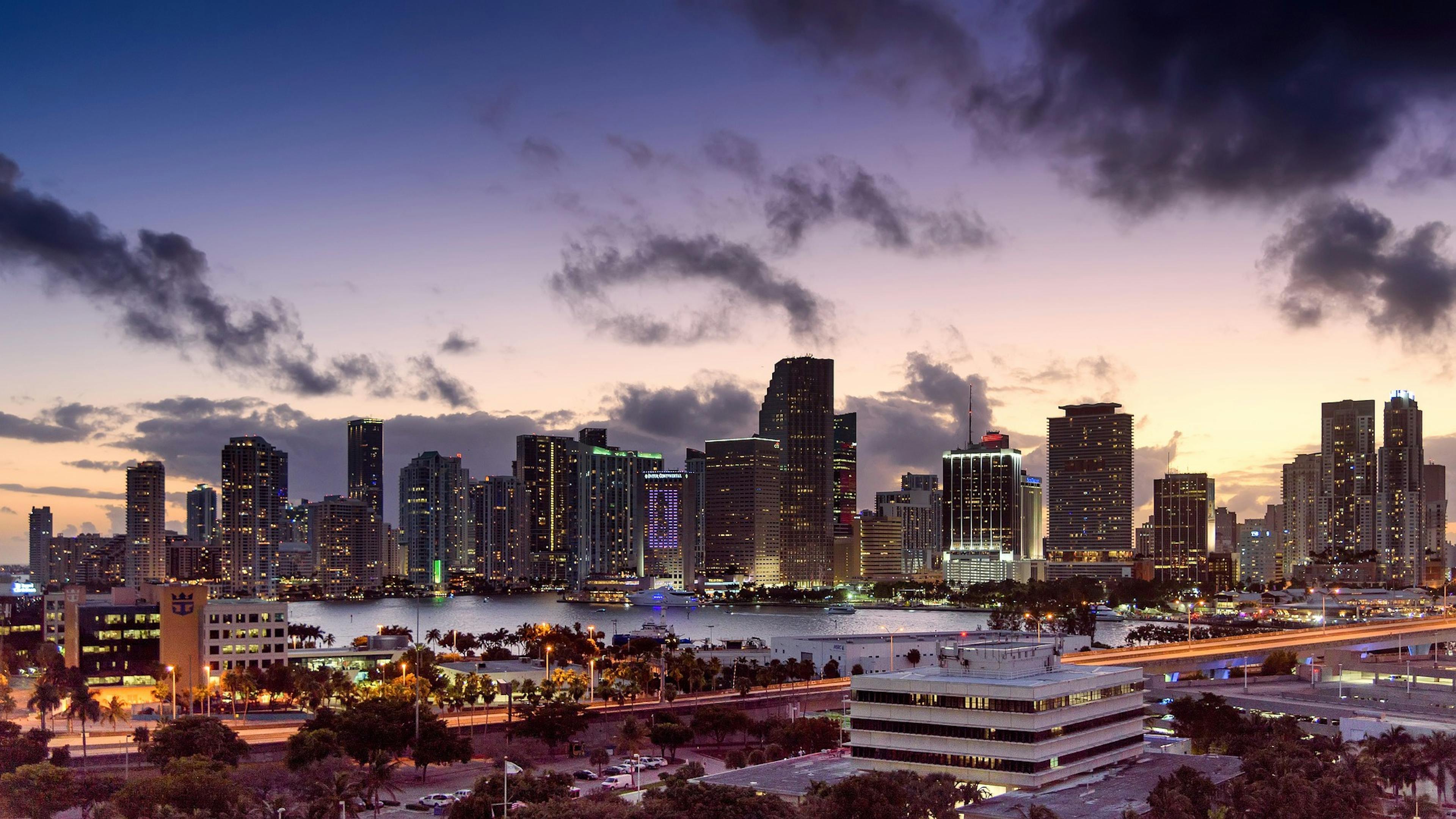 A long exposure wide view of Miami’s skyline at night with streaks of bright light from the highway in the foreground 
