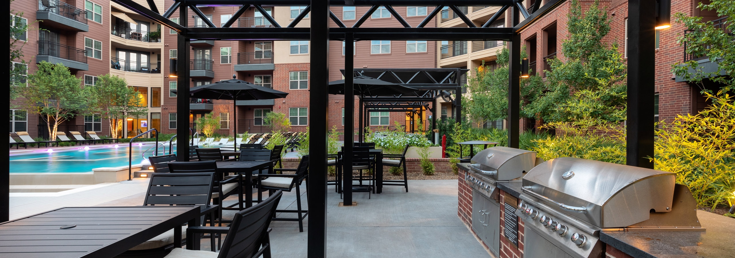 AMLI Grapevine poolside grill station with black chairs and tables surrounded by lush greenery and brick building exterior