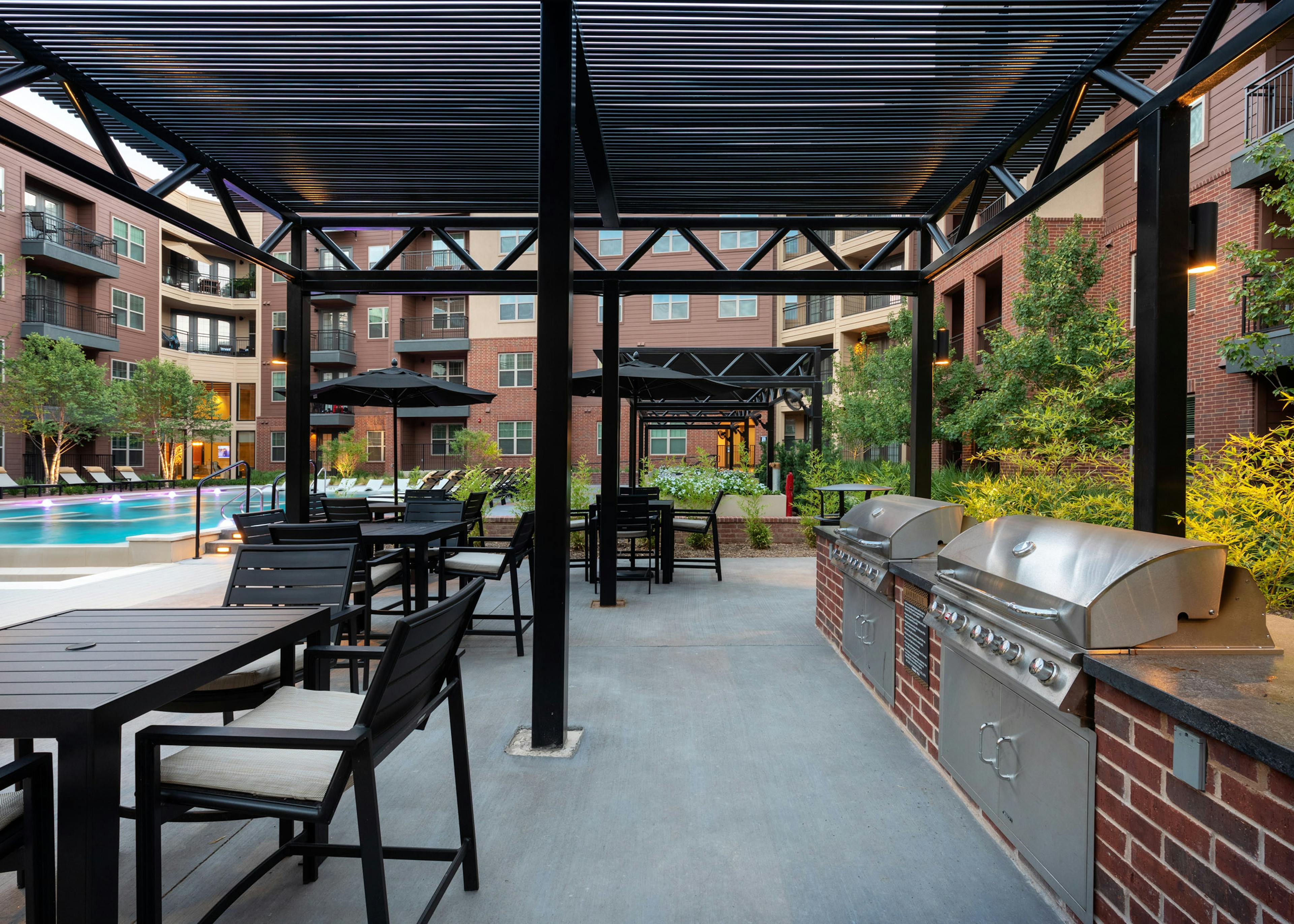 AMLI Grapevine poolside grill station with black chairs and tables surrounded by lush greenery and brick building exterior