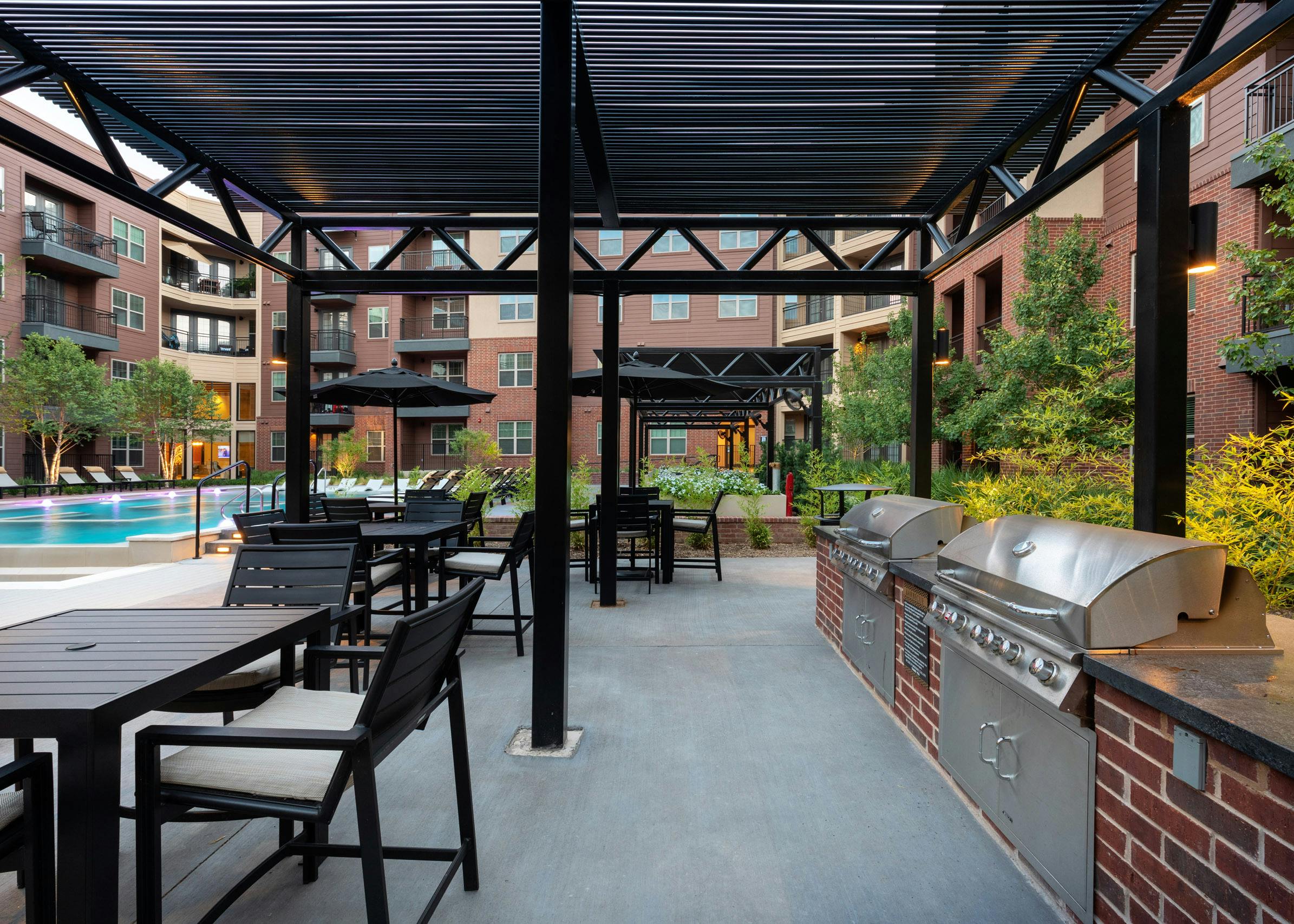 AMLI Grapevine poolside grill station with black chairs and tables surrounded by lush greenery and brick building exterior