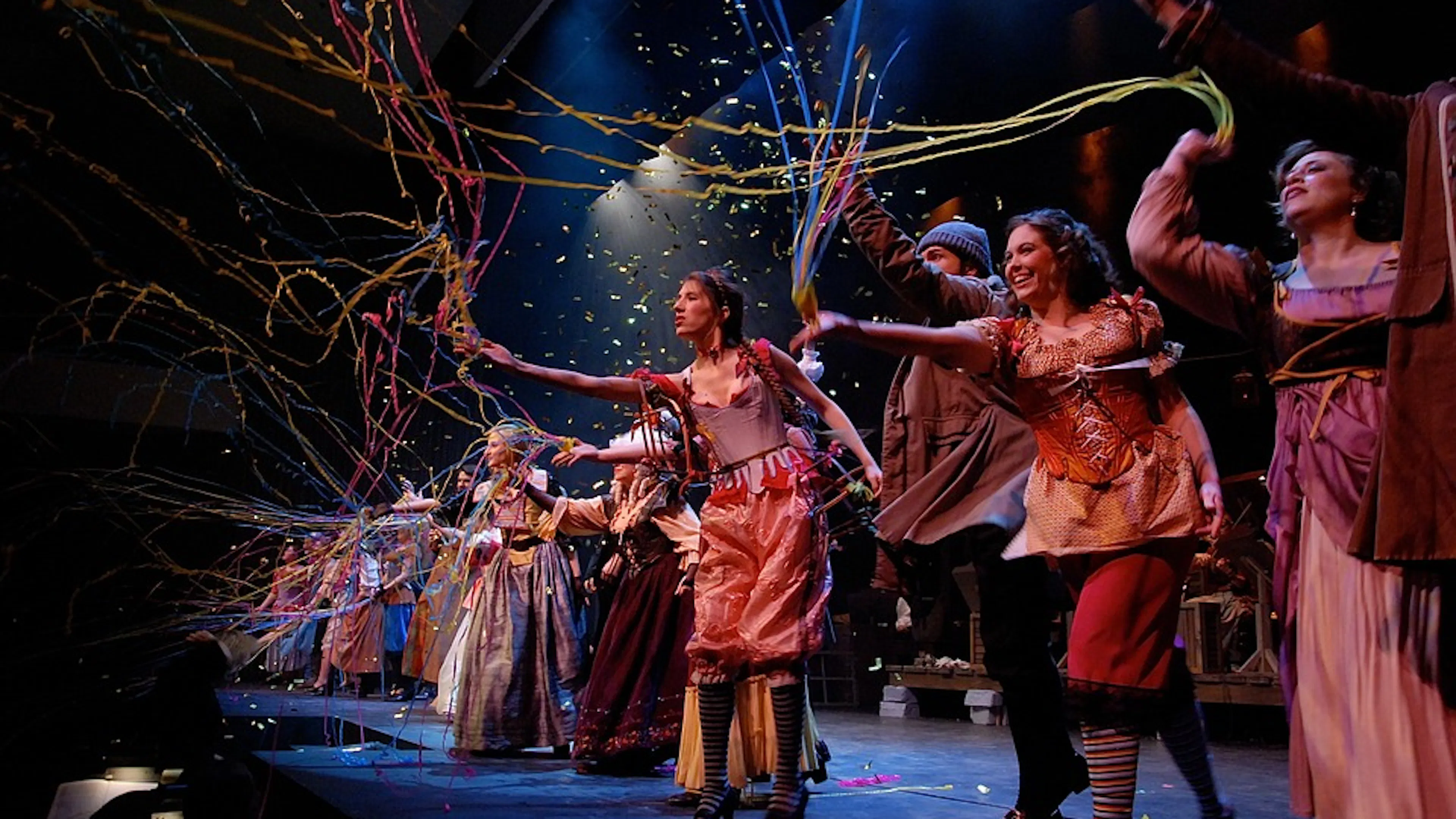 A theater stage with colorfully dressed actors at the edge of it throwing streamers out toward the crowd