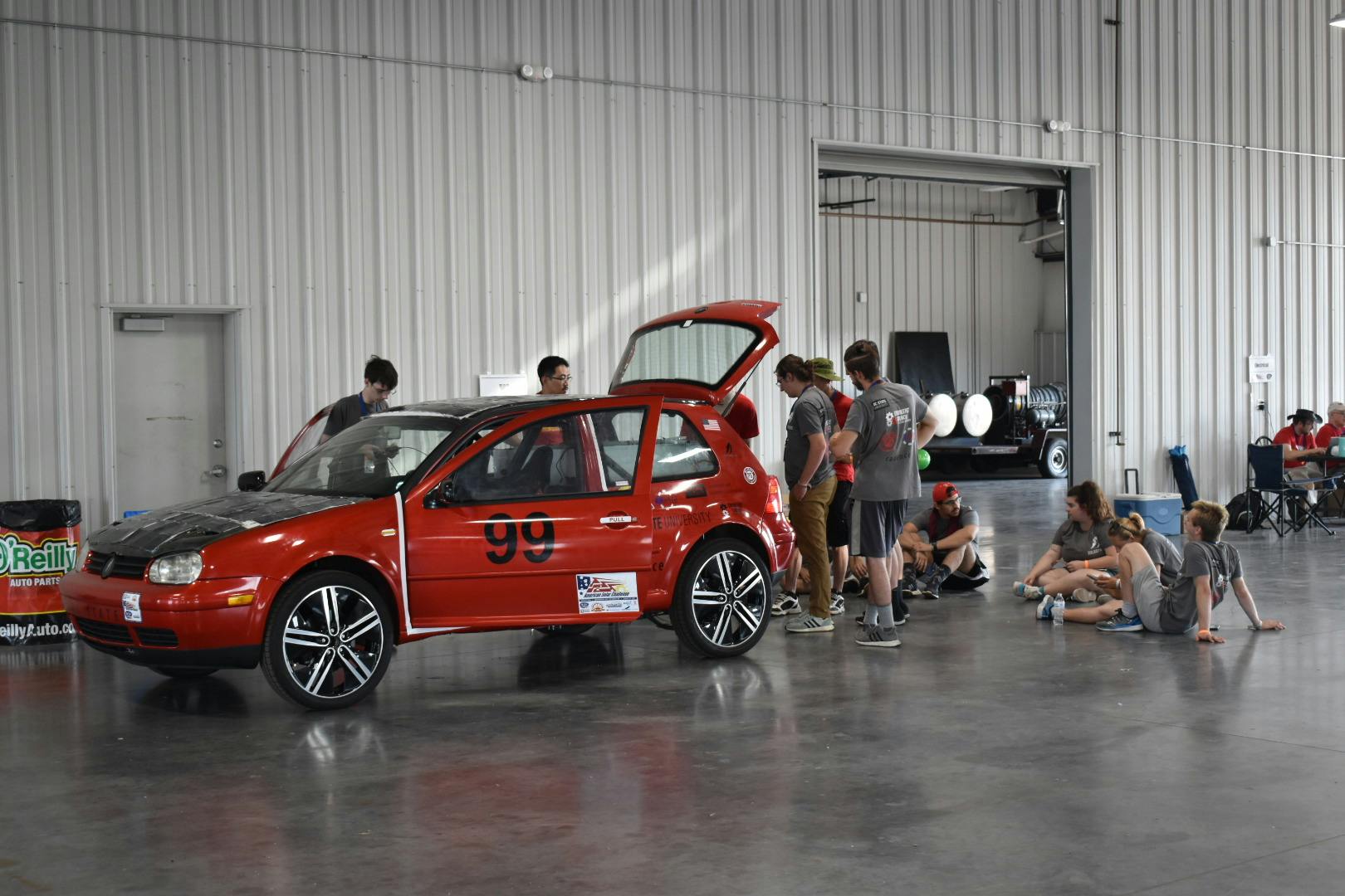 A red hatchback car covered in solar panels in a garage surrounded by a team of engineers working on it