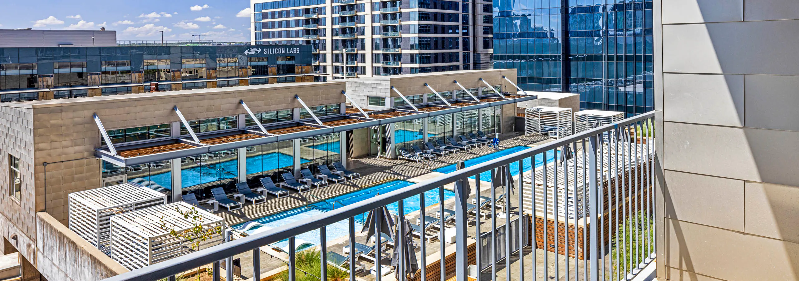 View of rooftop pool and lounge chairs from apartment balcony above at AMLI on 2nd with blue sky and city buildings