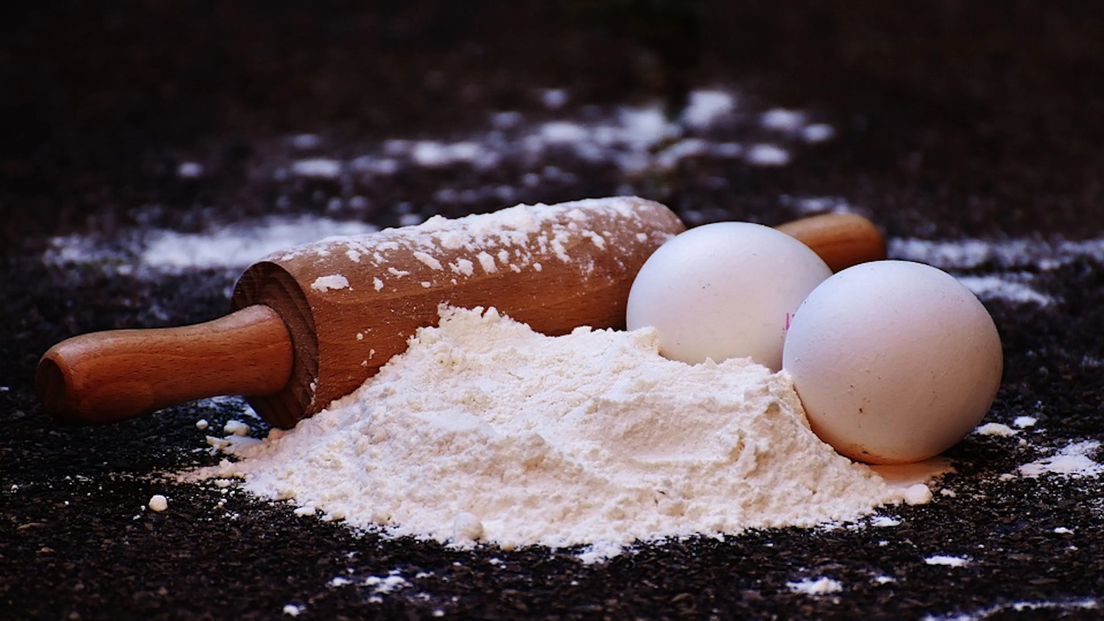 A little mound of flour on a dark table with a wooden rolling pin and two eggs beside it