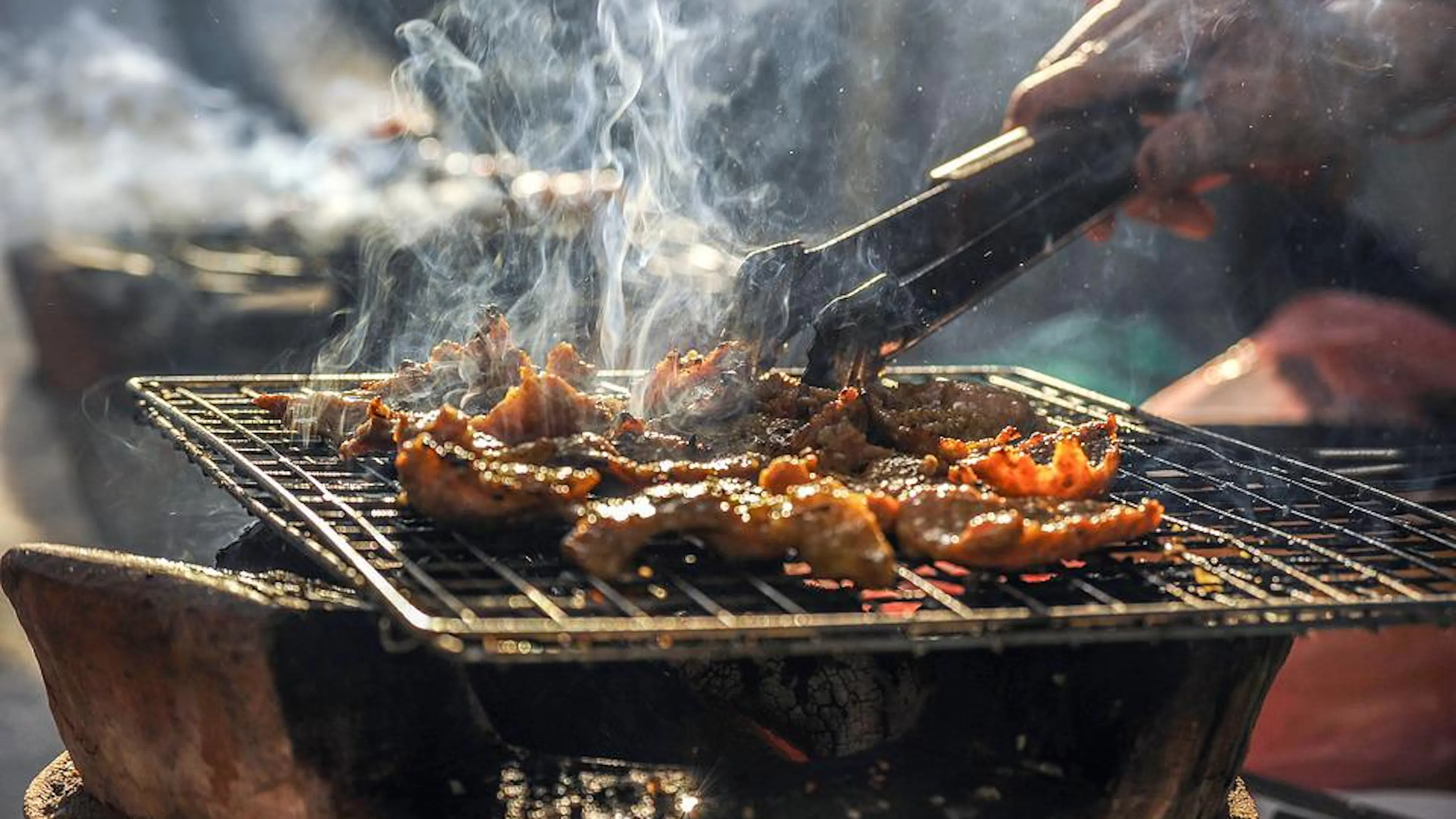 A metal grill over a charcoal fire with someone flipping meat with tongs. 
