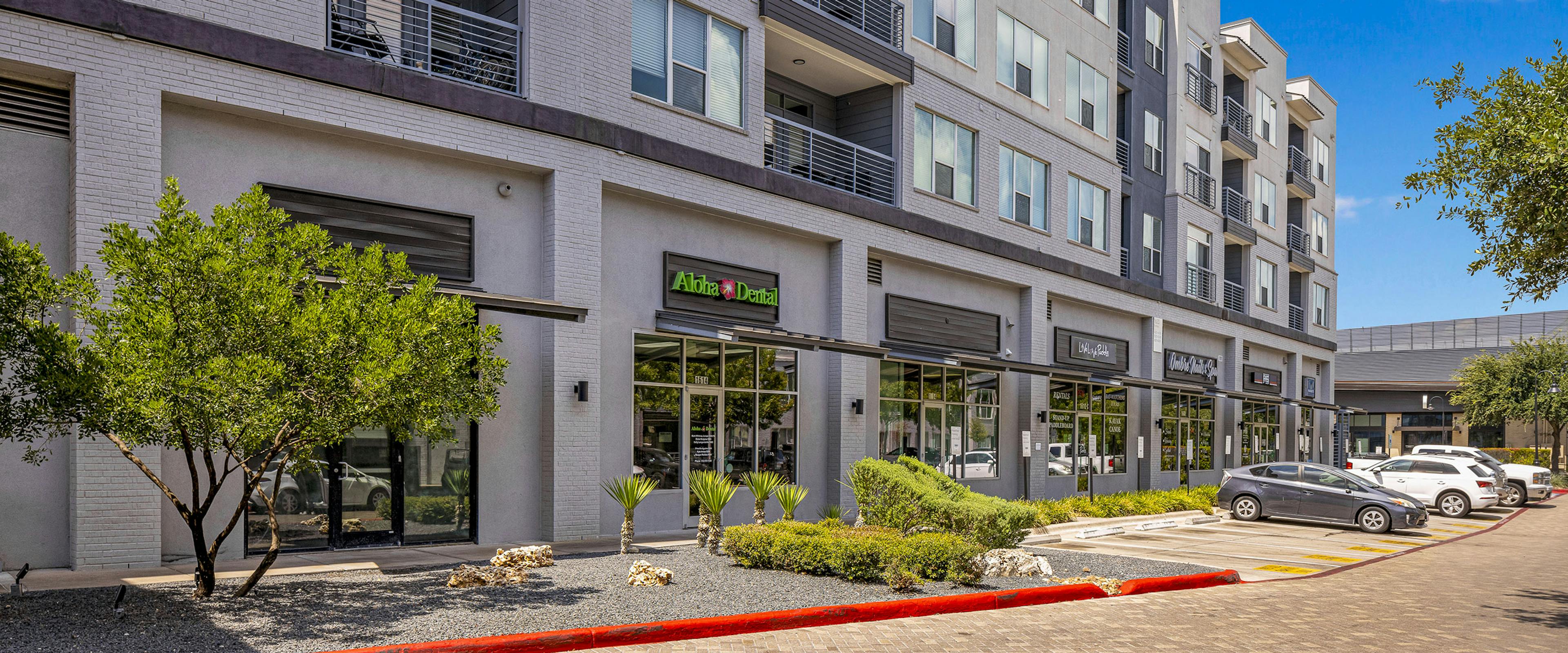 AMLI South Shore building exterior with gray façade and various storefronts with greenery and cars in front on brick street