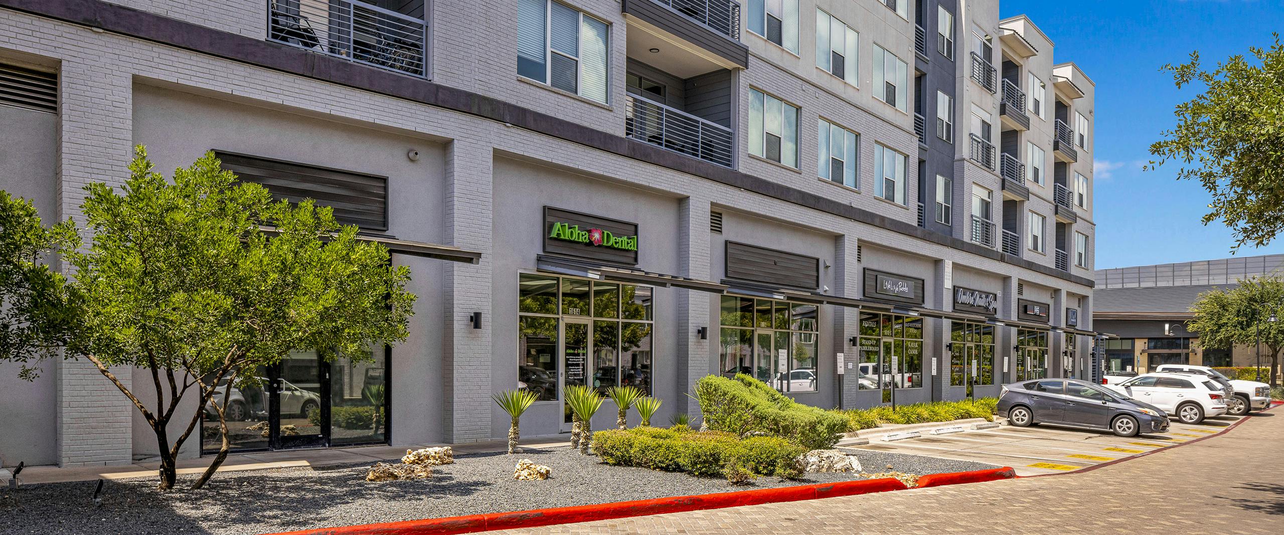 AMLI South Shore building exterior with gray façade and various storefronts with greenery and cars in front on brick street