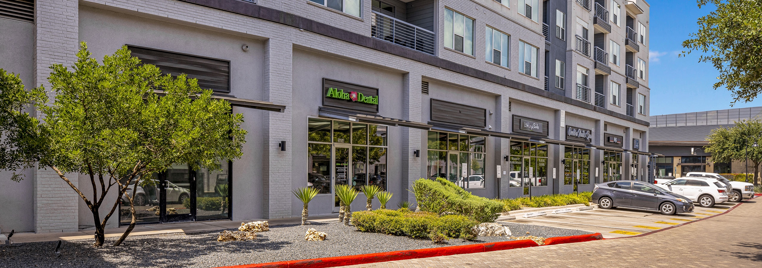 AMLI South Shore building exterior with gray façade and various storefronts with greenery and cars in front on brick street
