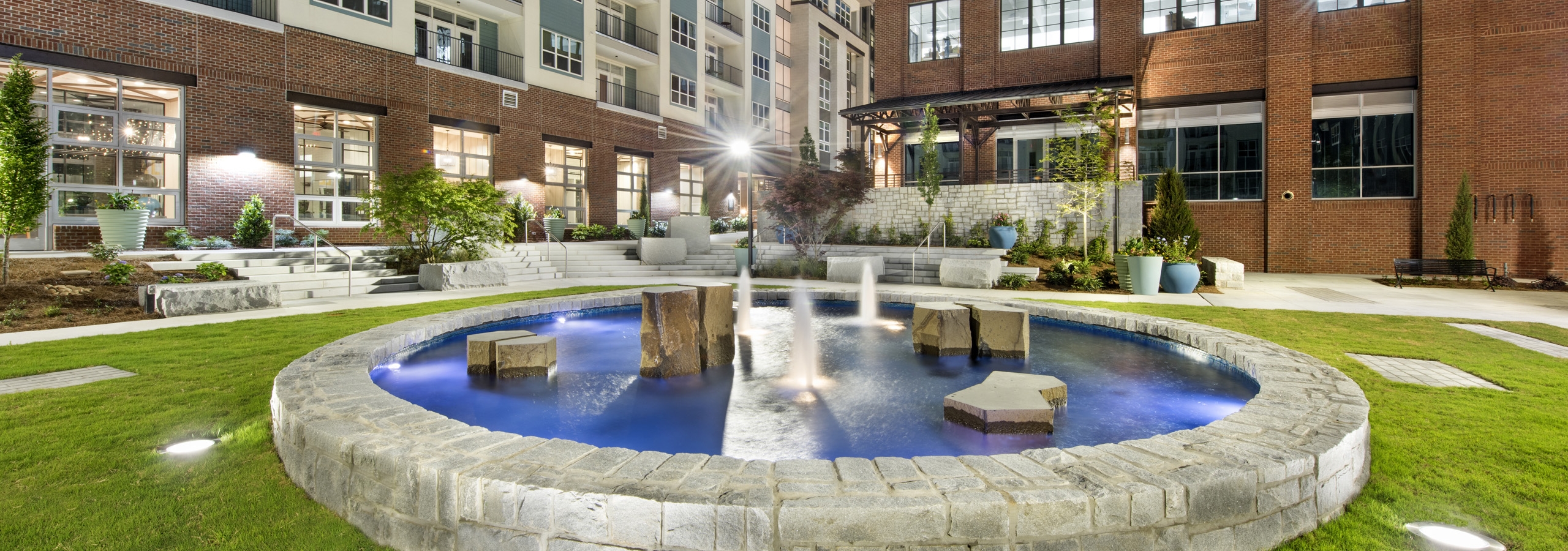 Nighttime view of the AMLI Decatur courtyard showcasing a lit stone fountain with blue water surrounded by manicured grass