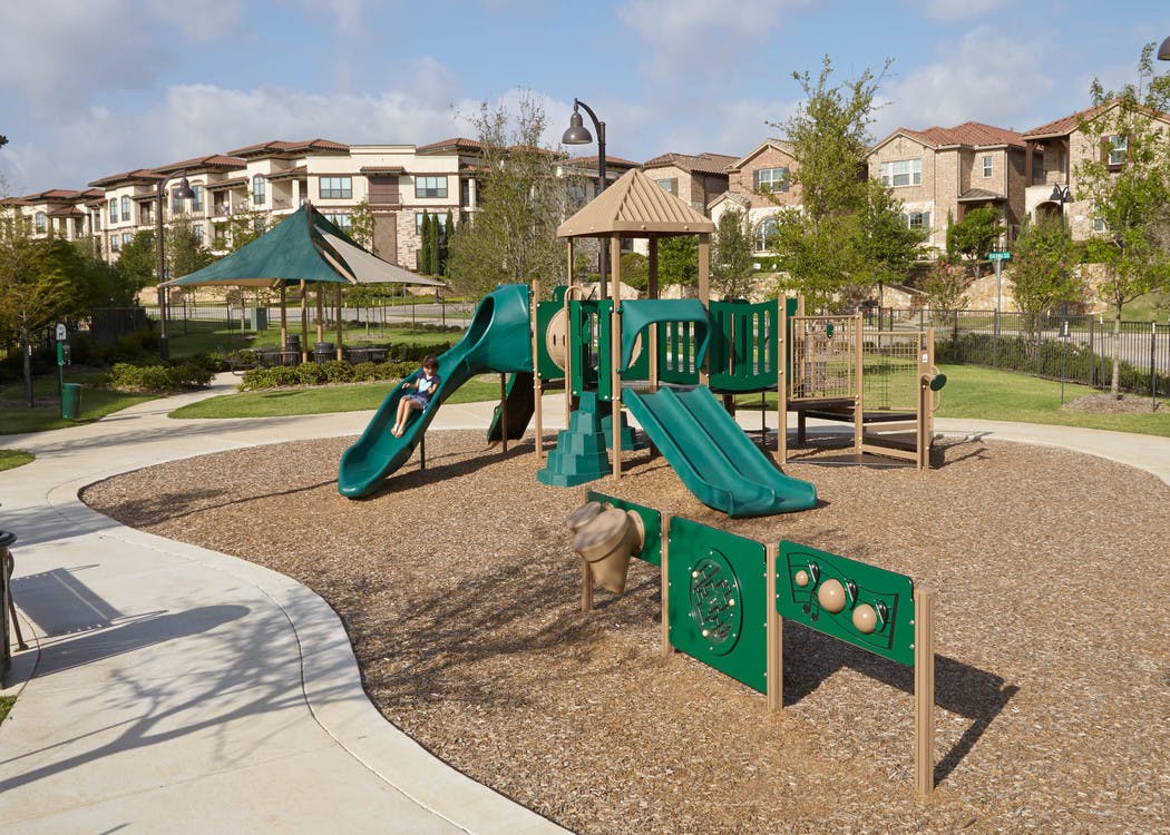 Playground with green slides on a bed of woodchips with a blue sky, trees and apartment building facade in the background