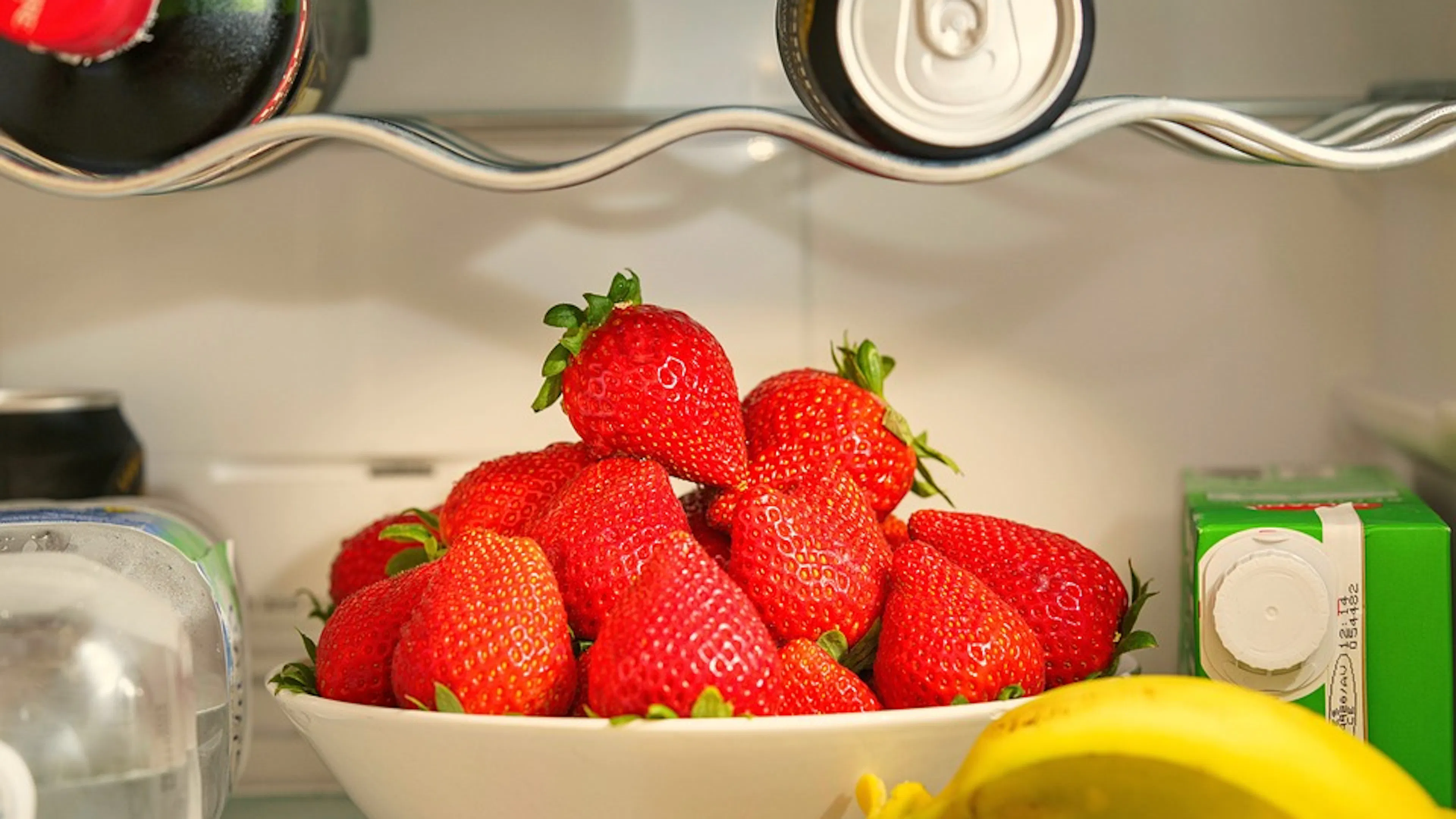 A bowl of strawberries on a shelf in the fridge with bananas and milk around it