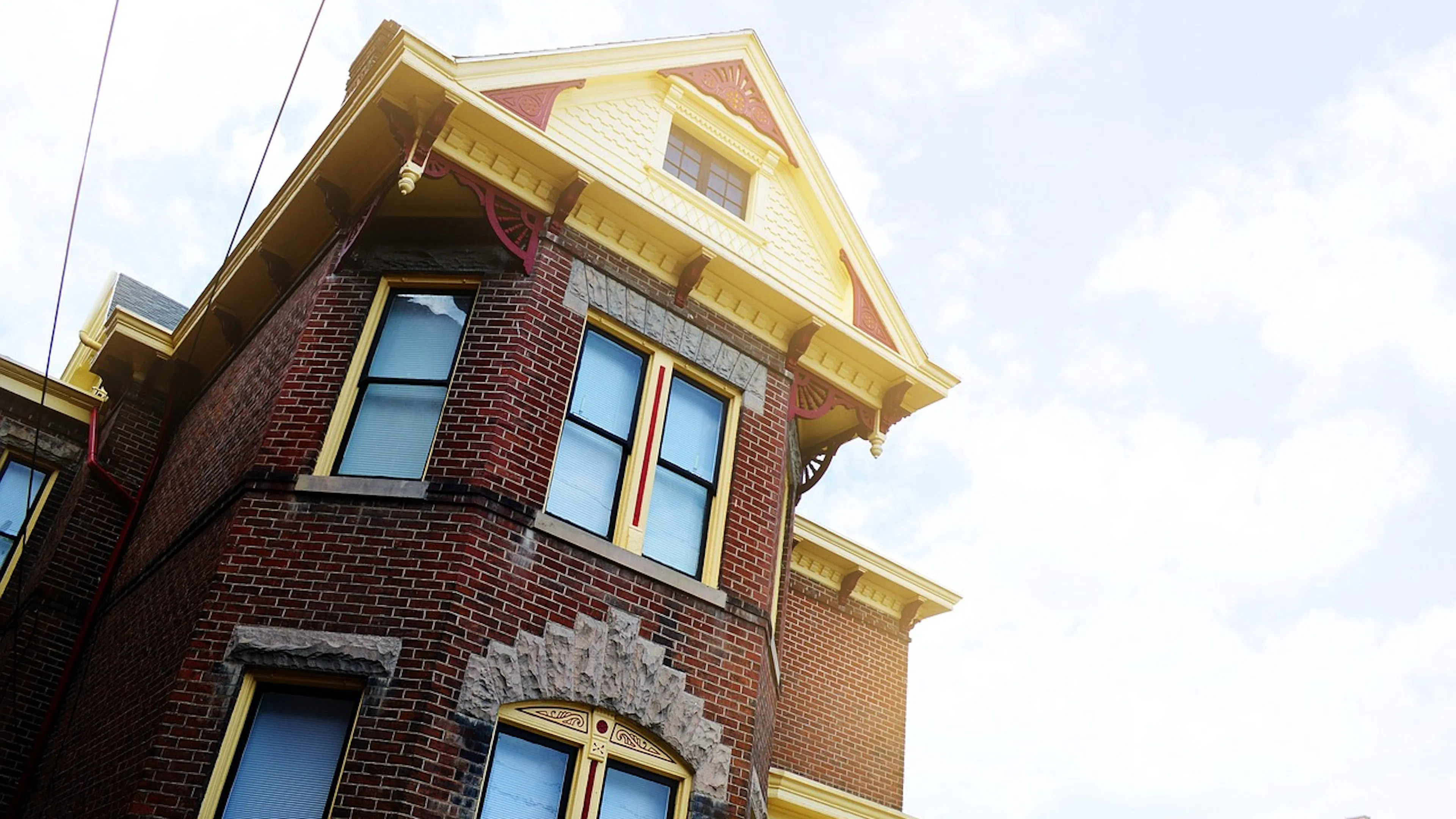 Facade of a three story brick mansion with yellow trim around the windows and roof with a blue and cloudy sky in the background
