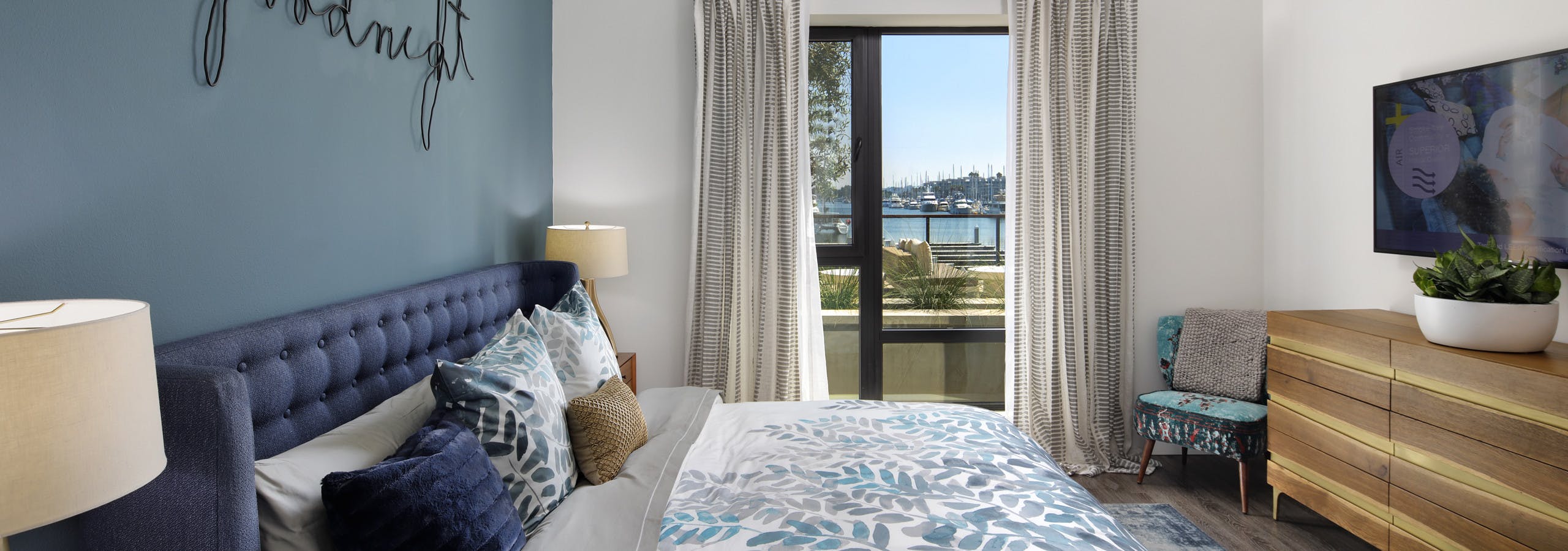 Interior view of AMLI Marina Del Rey bedroom with blue upholstered queen headboard, big screen TV and window facing marina