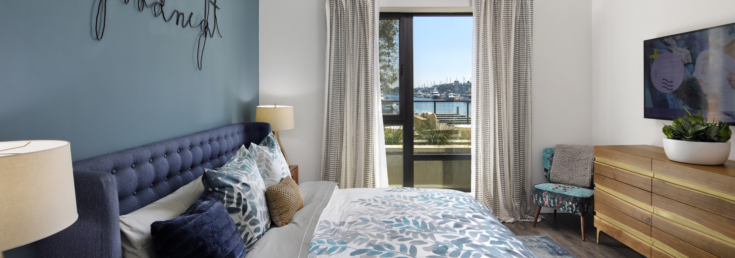 Interior view of AMLI Marina Del Rey bedroom with blue upholstered queen headboard, big screen TV and window facing marina