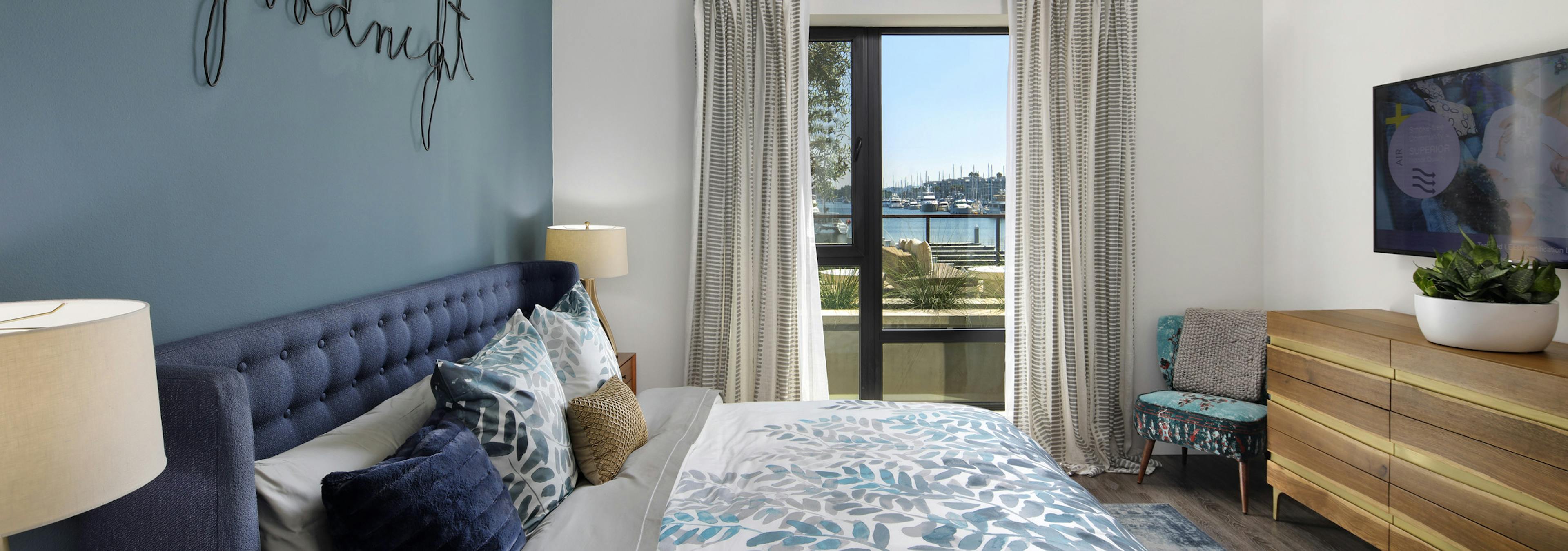 Interior view of AMLI Marina Del Rey bedroom with blue upholstered queen headboard, big screen TV and window facing marina