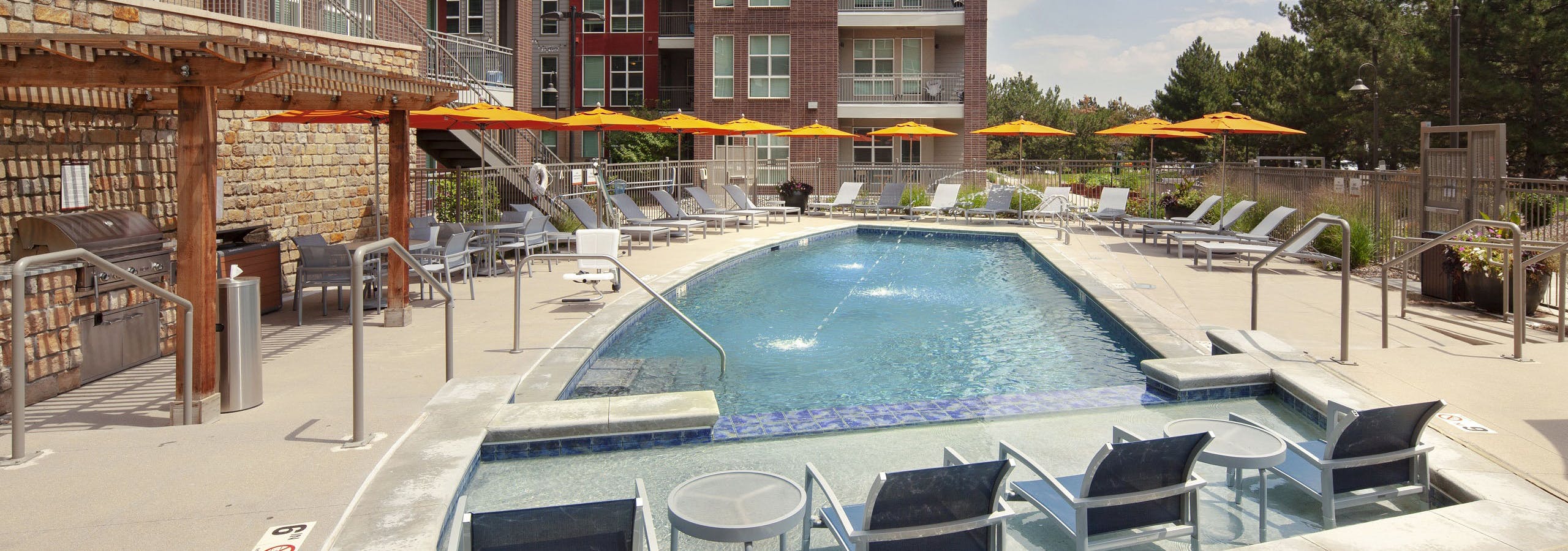 Daytime view at AMLI Dry Creek pool with lounge seating with tables and orange umbrellas and lush green trees and blue sky