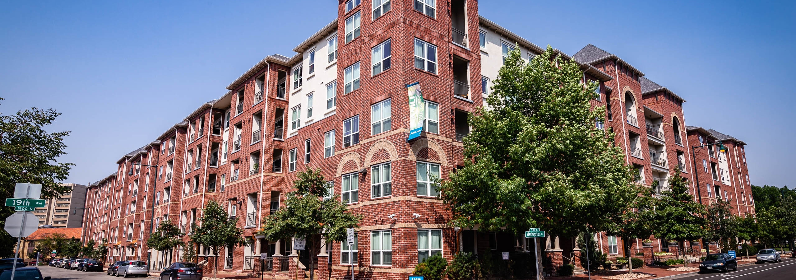Exterior view of AMLI Park Avenue apartment building with side streets and several cars park and large trees and blue sky