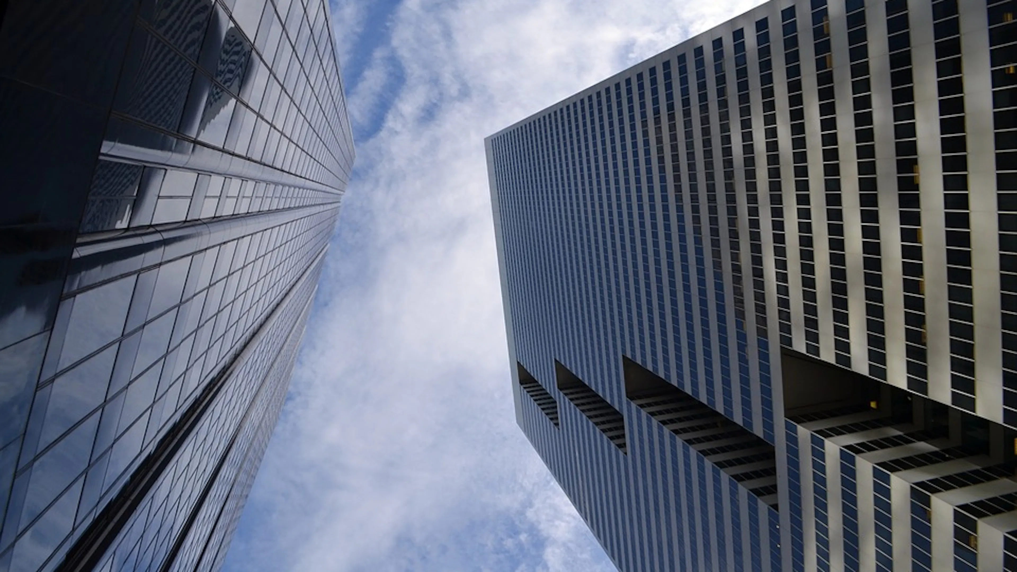 View of two glass covered skyscrapers looking straight up toward a cloudy blue sky