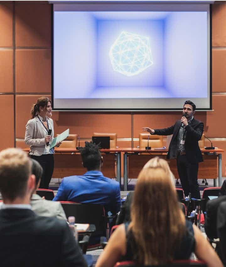 Two speakers on stage before a large screen at a conference presenting to an audience in an auditorium