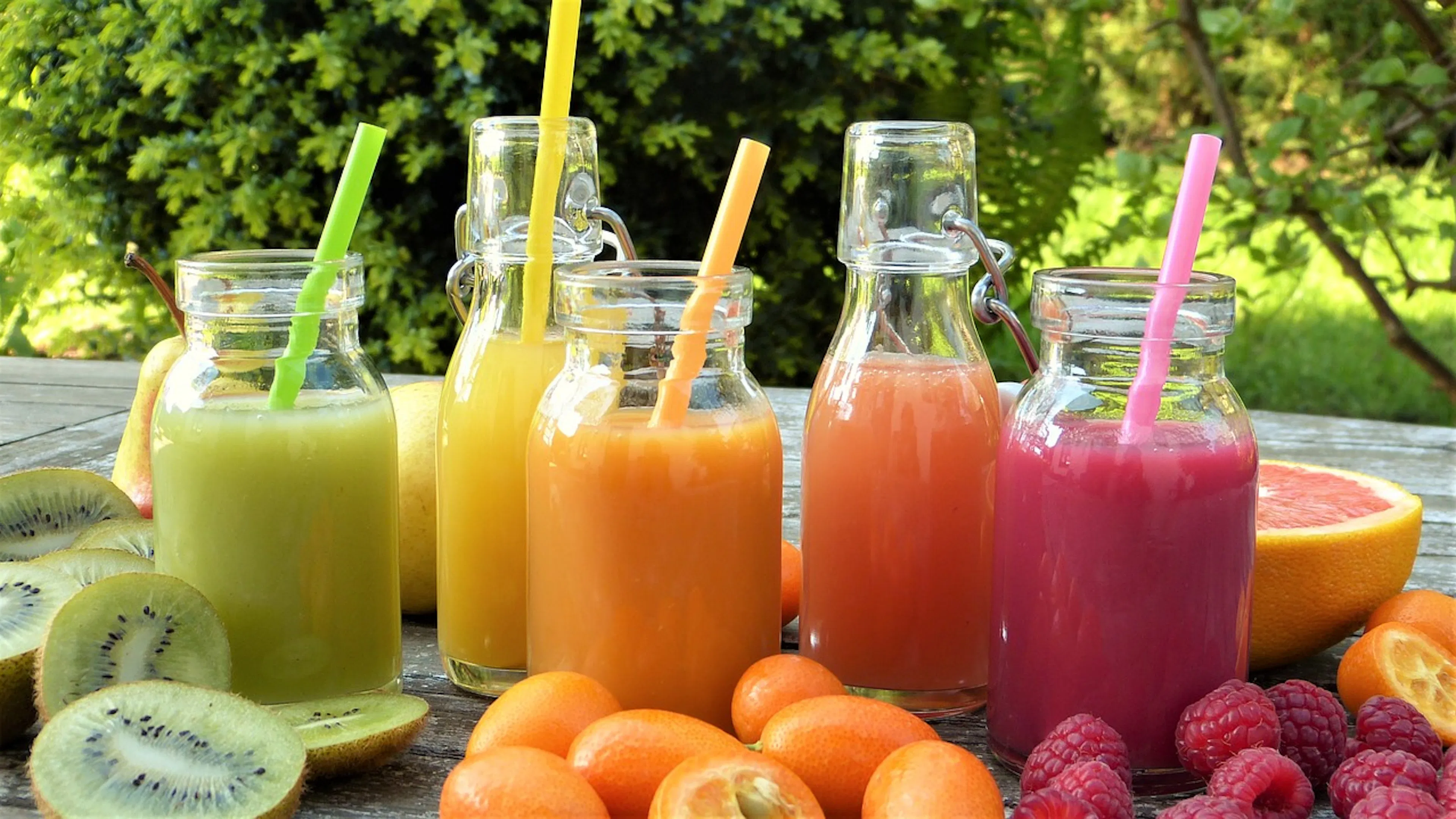 Five glass jars with straws on a wooden table all filled with various colored juices and surrounded by colorful fruits like kiwis and raspberries