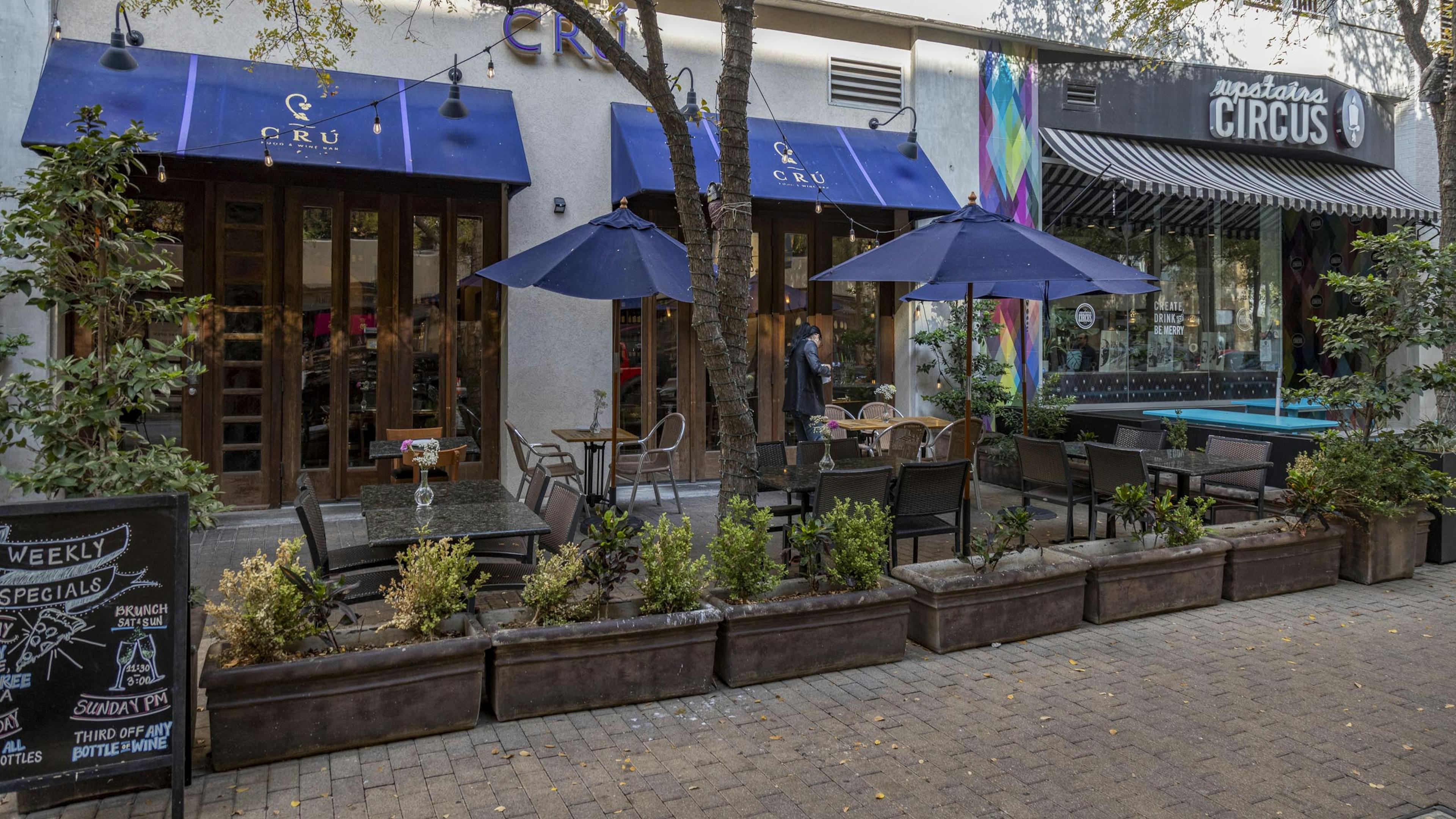 Daytime photo of Cru Wine Bar exterior with landscaped plants and outdoor seating shaded by blue umbrellas