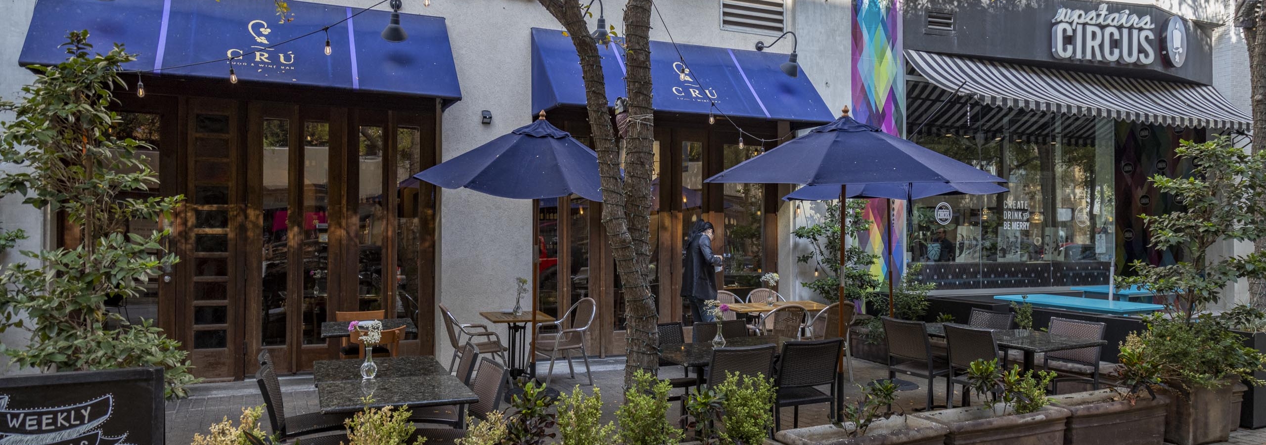 Daytime photo of Cru Wine Bar exterior with landscaped plants and outdoor seating shaded by blue umbrellas