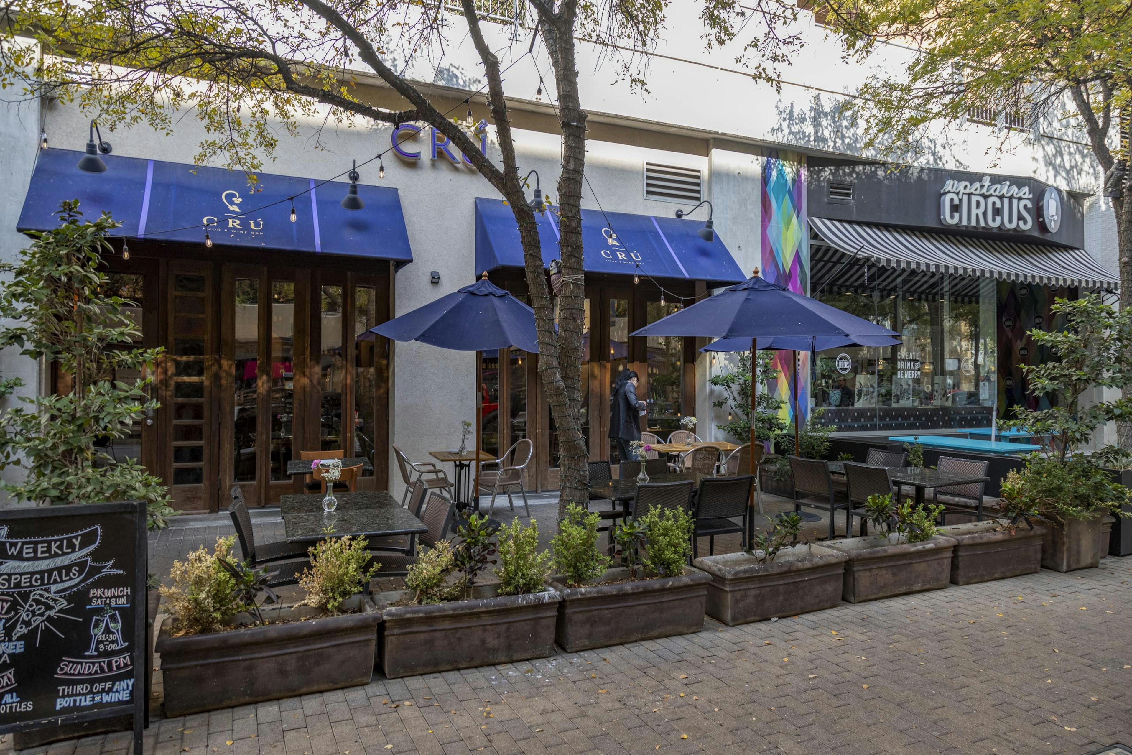 Daytime photo of Cru Wine Bar exterior with landscaped plants and outdoor seating shaded by blue umbrellas