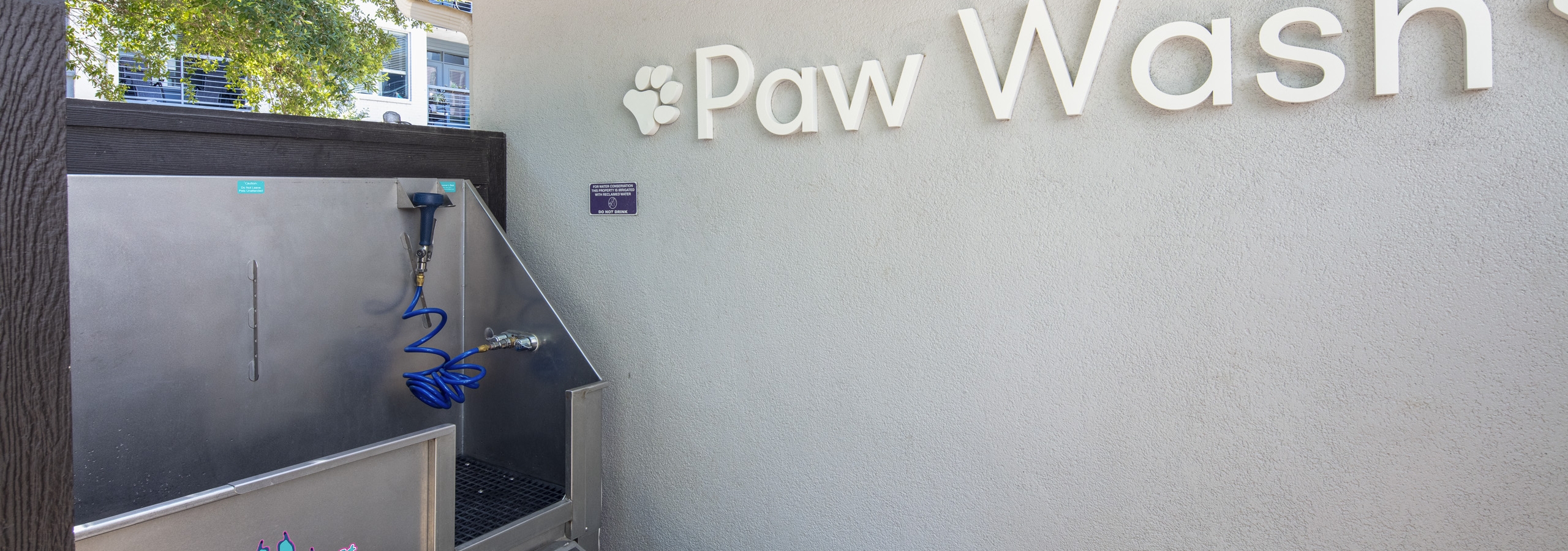 AMLI South Shore apartments outdoor paw wash with gray wall and a stainless washing tub with blue sprayer with trees in rear