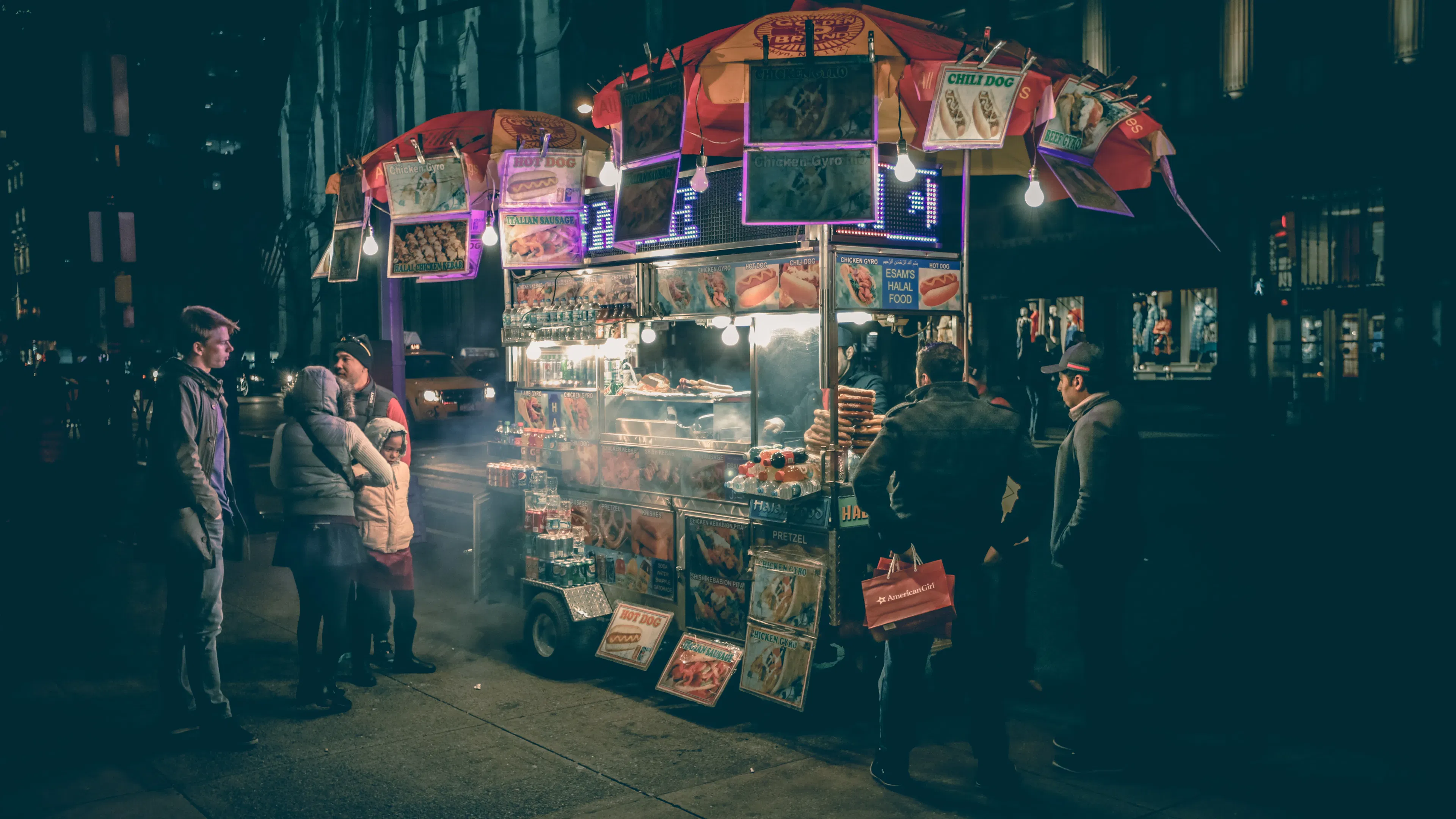 A dimly lit hot dog stand with a few people waiting in line and some smoke wafting out of the front