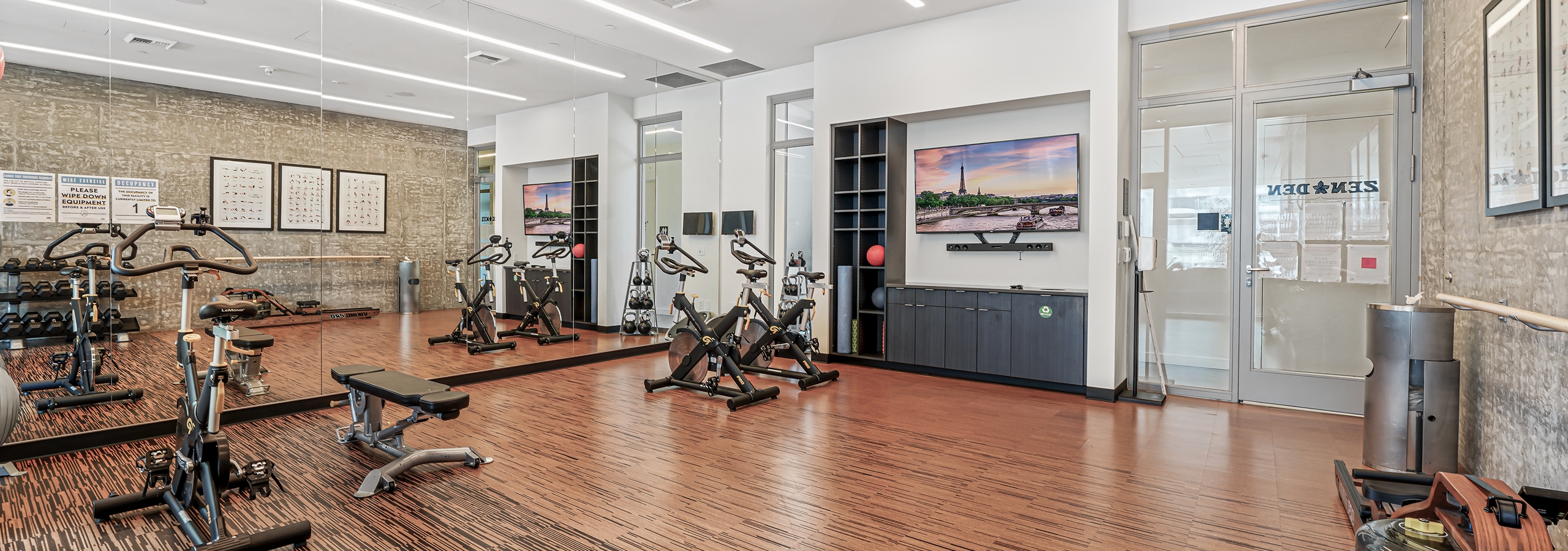 AMLI Arc Zen Den with spin bikes and weightlifting bench on wood grain floor and dumbbells and TV mounted over dark cabinet