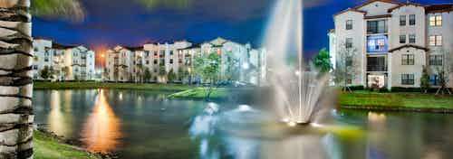 Nighttime exterior view of AMLI Doral apartment buildings surrounding a lake with a water fountain in the center