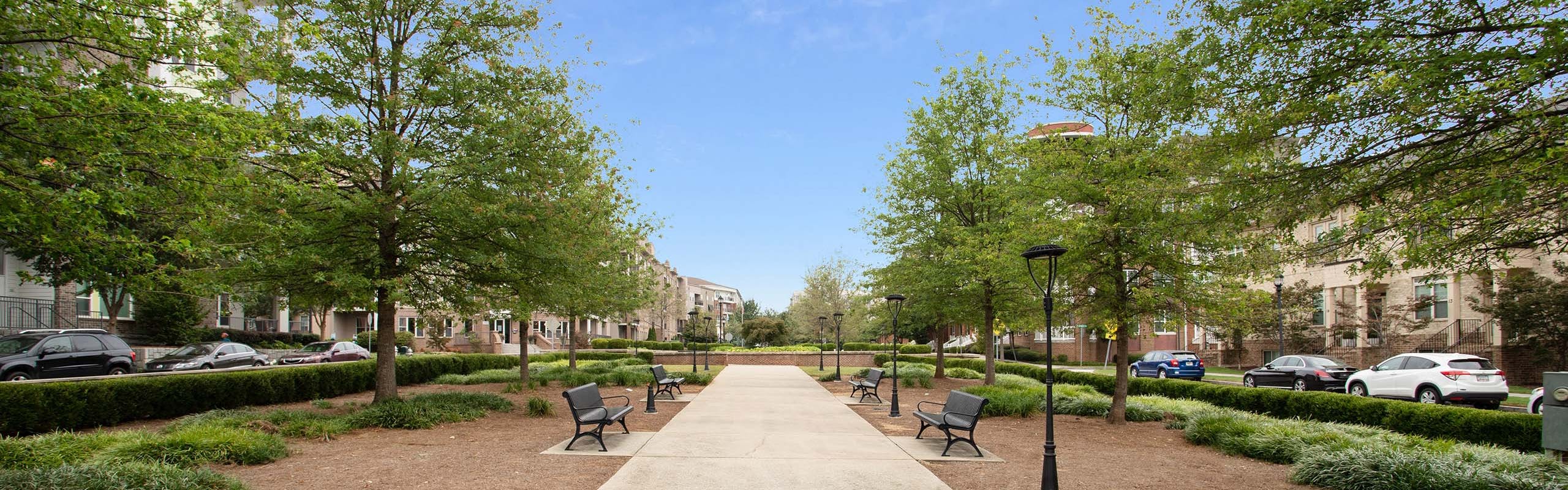 Daytime view of lush park walkway with alternating benches and lantern posts leading toward AMLI Lindbergh apartments