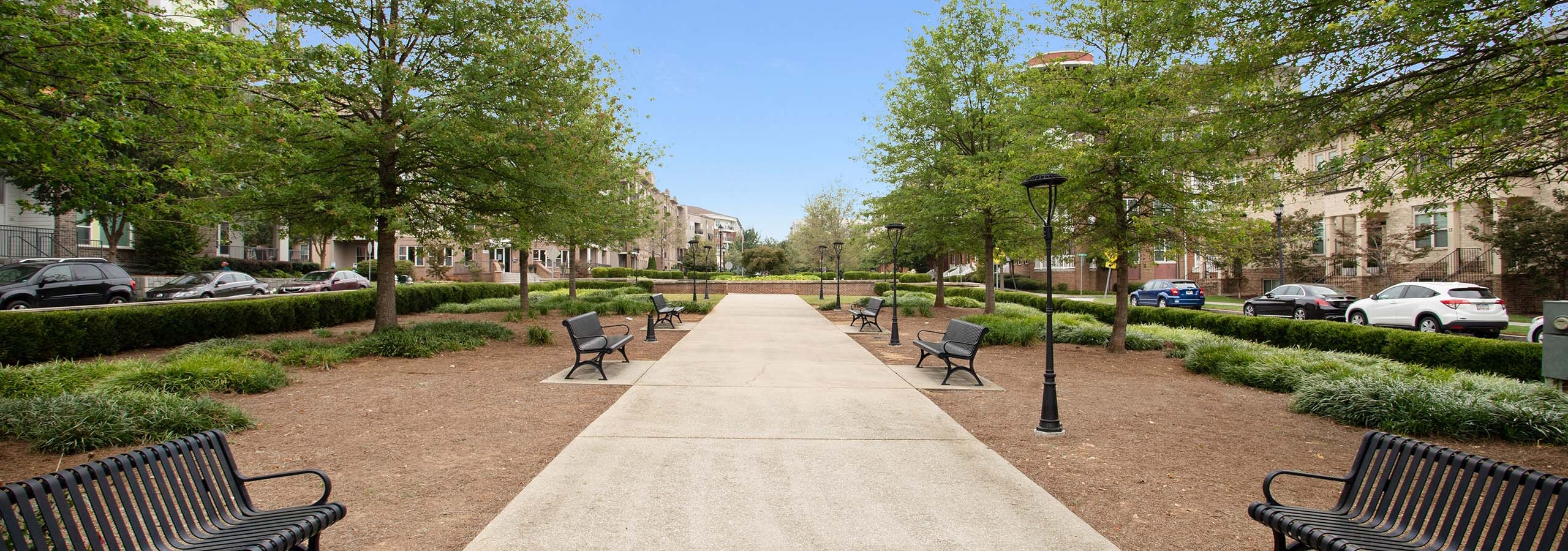 Daytime view of lush park walkway with alternating benches and lantern posts leading toward AMLI Lindbergh apartments