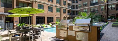 Daytime view of poolside gas grill station and tables with chairs and green umbrellas at AMLI Uptown apartments