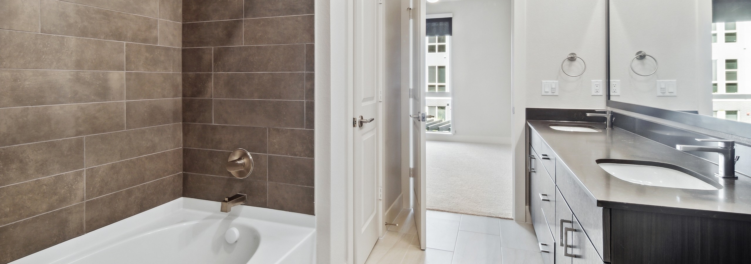AMLI RiNo bathroom with white garden tub with brown tile surround and dark vanity and countertop with silver finish hardware