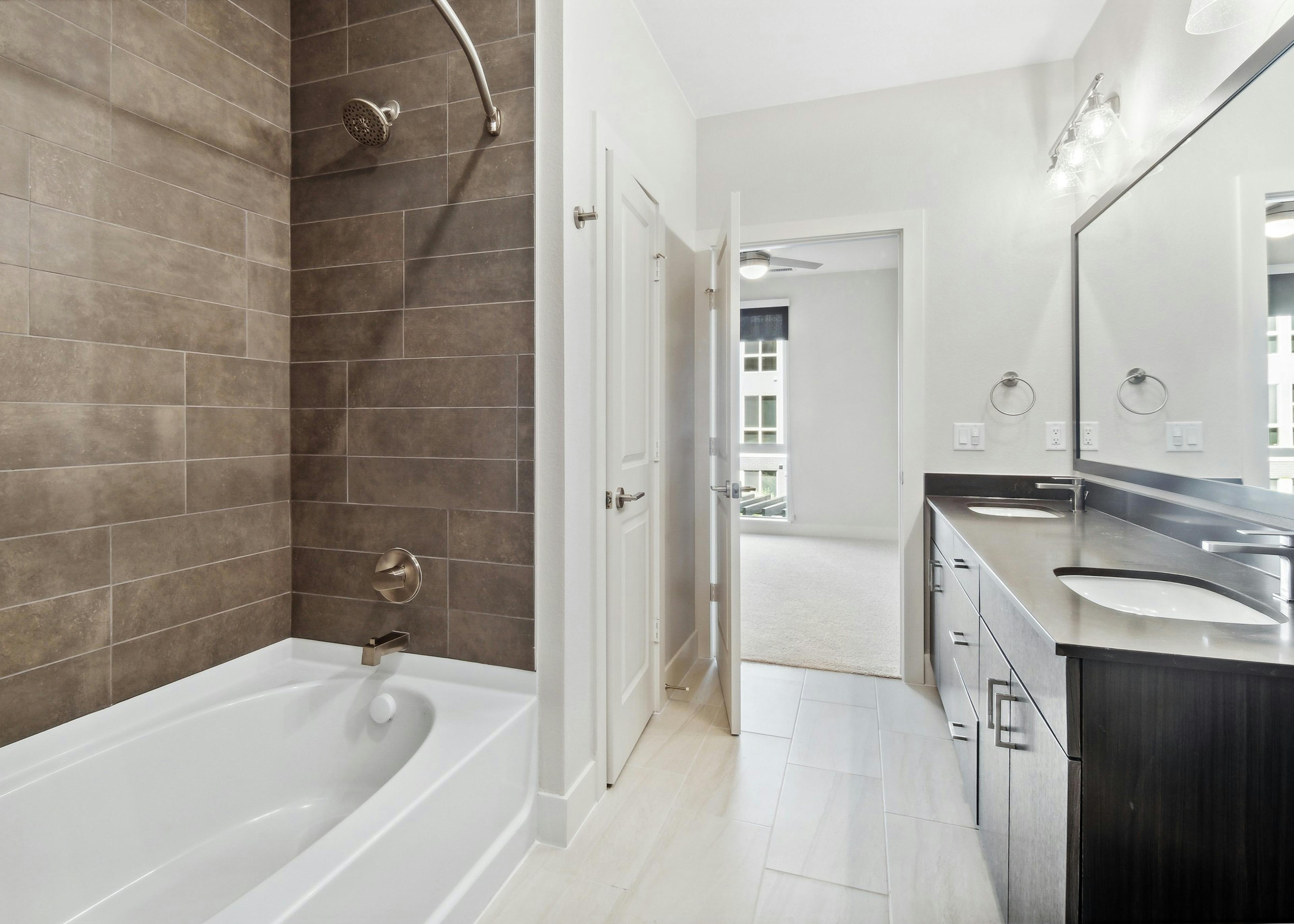 AMLI RiNo bathroom with white garden tub with brown tile surround and dark vanity and countertop with silver finish hardware