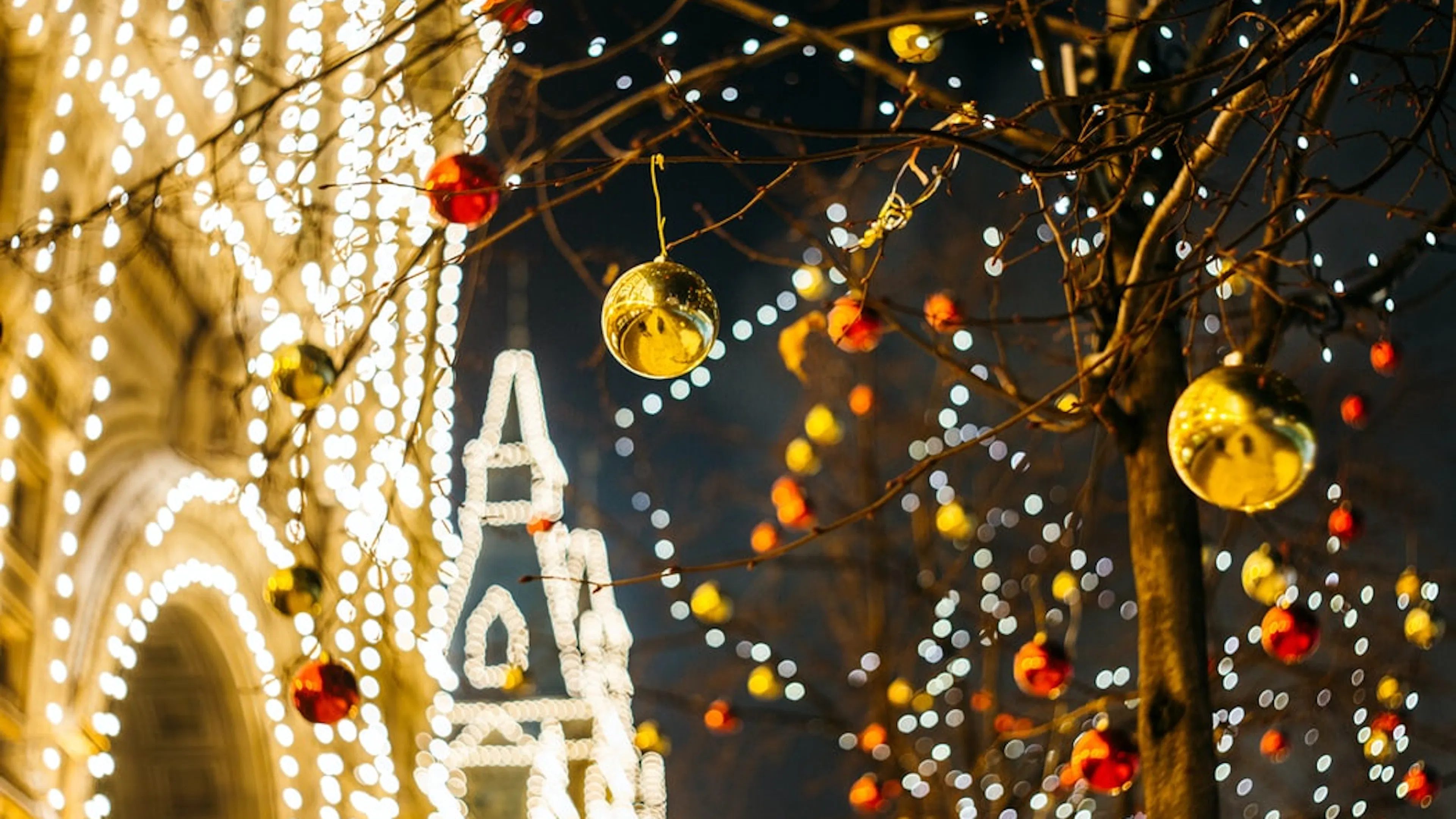 A close up of gold and red Christmas ornaments hanging from an outdoor tree and a building covered in string lights in the distance